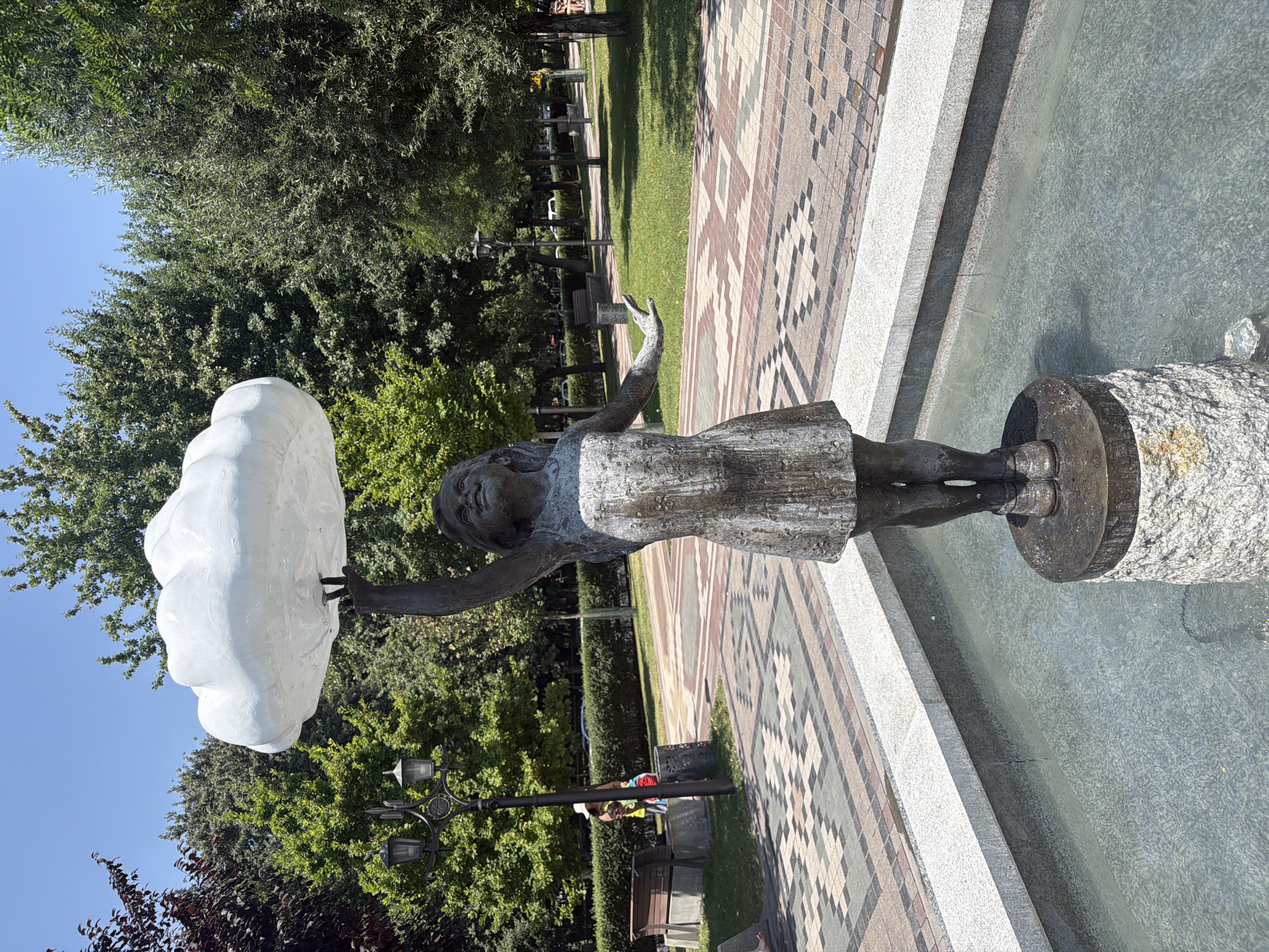 A bronze statue of a young girl holding a large white cloud above her head is depicted in a park fountain in Yerevan, Armenia. The girl is wearing a simple dress and shoes. The fountain has a stone base, and the water in the fountain is clear. The background shows lush green trees, a paved walkway with a geometric pattern, and a lamppost. In the distance, there are park benches, more trees, and a hint of a street with cars. The sky is clear and blue. The overall scene is tranquil and suggests a public park setting.