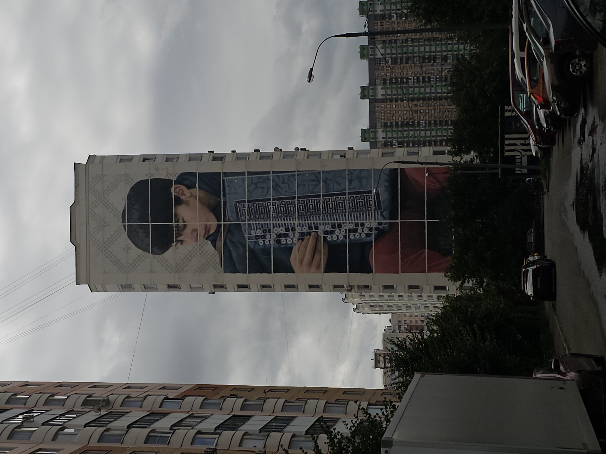 This is a medium shot of a residential building in Odintsovo, Russia, featuring a large mural of a young boy playing an accordion. The building is tall and slender, with a light-colored facade adorned with a traditional geometric pattern above the mural. The mural itself is realistic, depicting the boy from the chest up, with his head turned down, eyes closed, and hands on the accordion. The accordion is depicted in blue and silver with intricate detailing. The background is a cloudy, overcast sky. To the left of the muraled building is another, taller residential building with balconies. In the foreground, a street is visible with parked cars, including a large white truck on the left. Further in the background, more residential buildings of varying heights and colors are visible, along with trees and a street lamp. The overall scene suggests an urban residential environment on a cloudy day.
