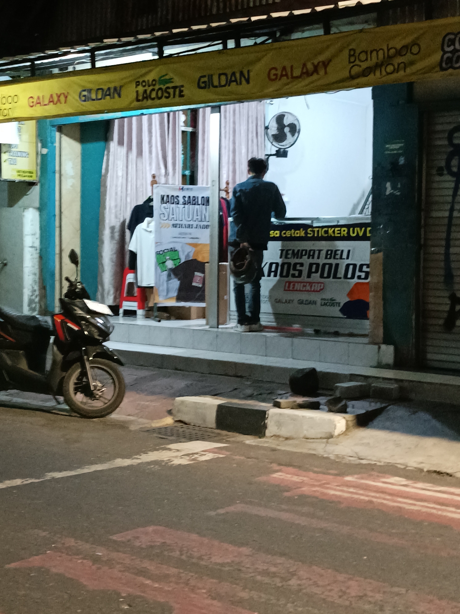 The image depicts a storefront in Yogyakarta, Indonesia, at night. A yellow banner with various brand names like "GILDAN," "LACOSTE," and "GALAXY" is visible above the entrance. Inside the store, a person is standing with their back to the camera, wearing a dark blue shirt and black pants, holding a bag. Various garments, including t-shirts and polo shirts, are displayed on racks and stands. A sign on display reads "KAOS SABLON SATUAN" (Custom T-Shirt Printing) and "TEMPAT BELI KAOS POLOS" (Place to Buy Plain T-Shirts). Outside the store, a scooter is parked on the side of the road. The street in front of the store has a pedestrian crossing painted in red and white. The overall scene suggests a commercial establishment operating in a typical urban environment.