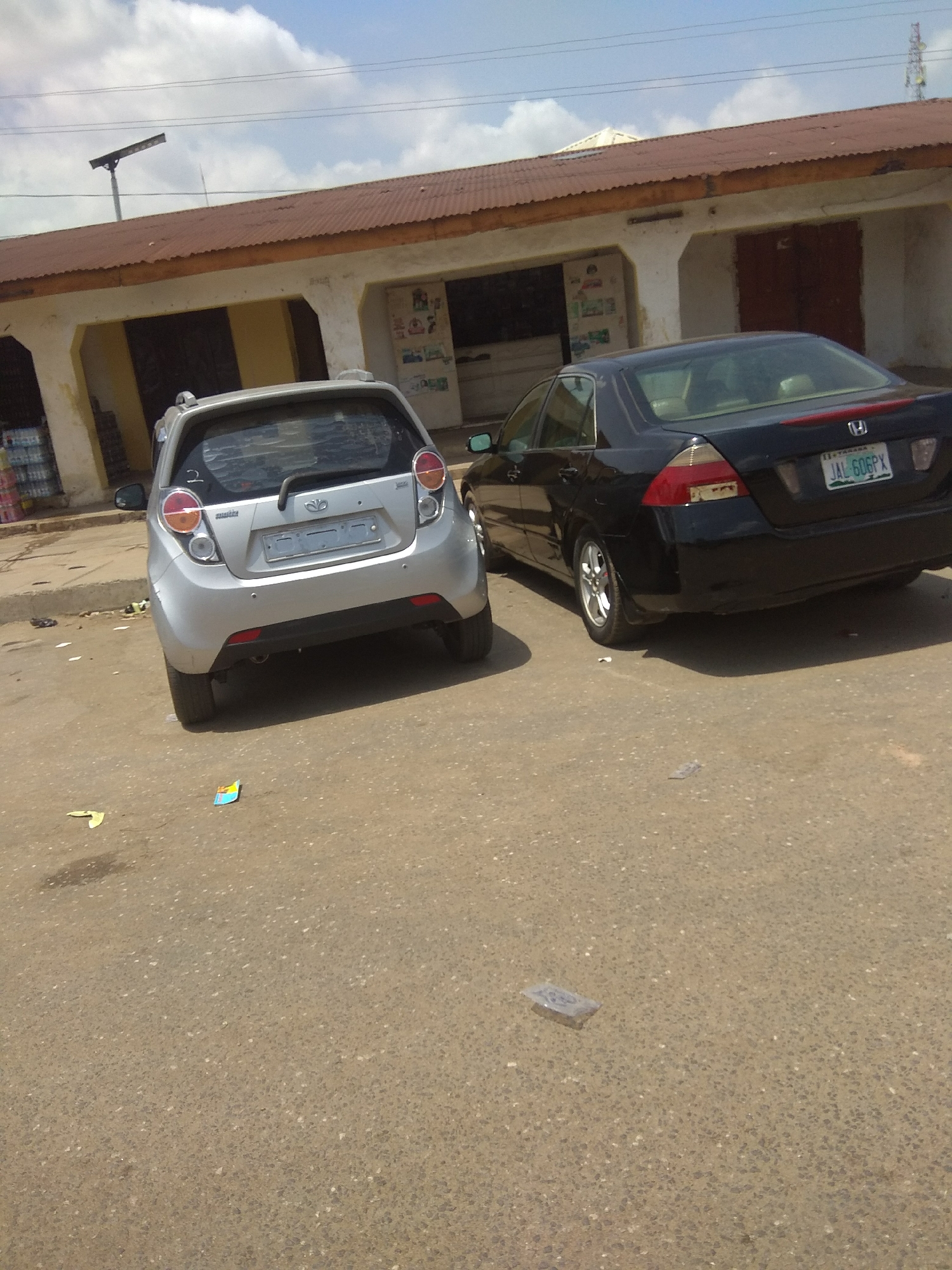 Two cars are parked on a paved surface in front of a building with a corrugated metal roof. The car on the left is a small, silver hatchback, likely a Daewoo Matiz, with its rear facing the camera. The car on the right is a black sedan, possibly a Honda Accord, with its rear also facing the camera. The license plate of the black sedan is visible and reads "JAL 606PX". The building behind the cars appears to be a shop or commercial establishment with closed doors and windows. The ground is a rough asphalt or concrete surface, with some debris and scattered papers visible. The sky above is partly cloudy. The scene is likely located in Jalingo, Nigeria, based on the context provided by the prompt.