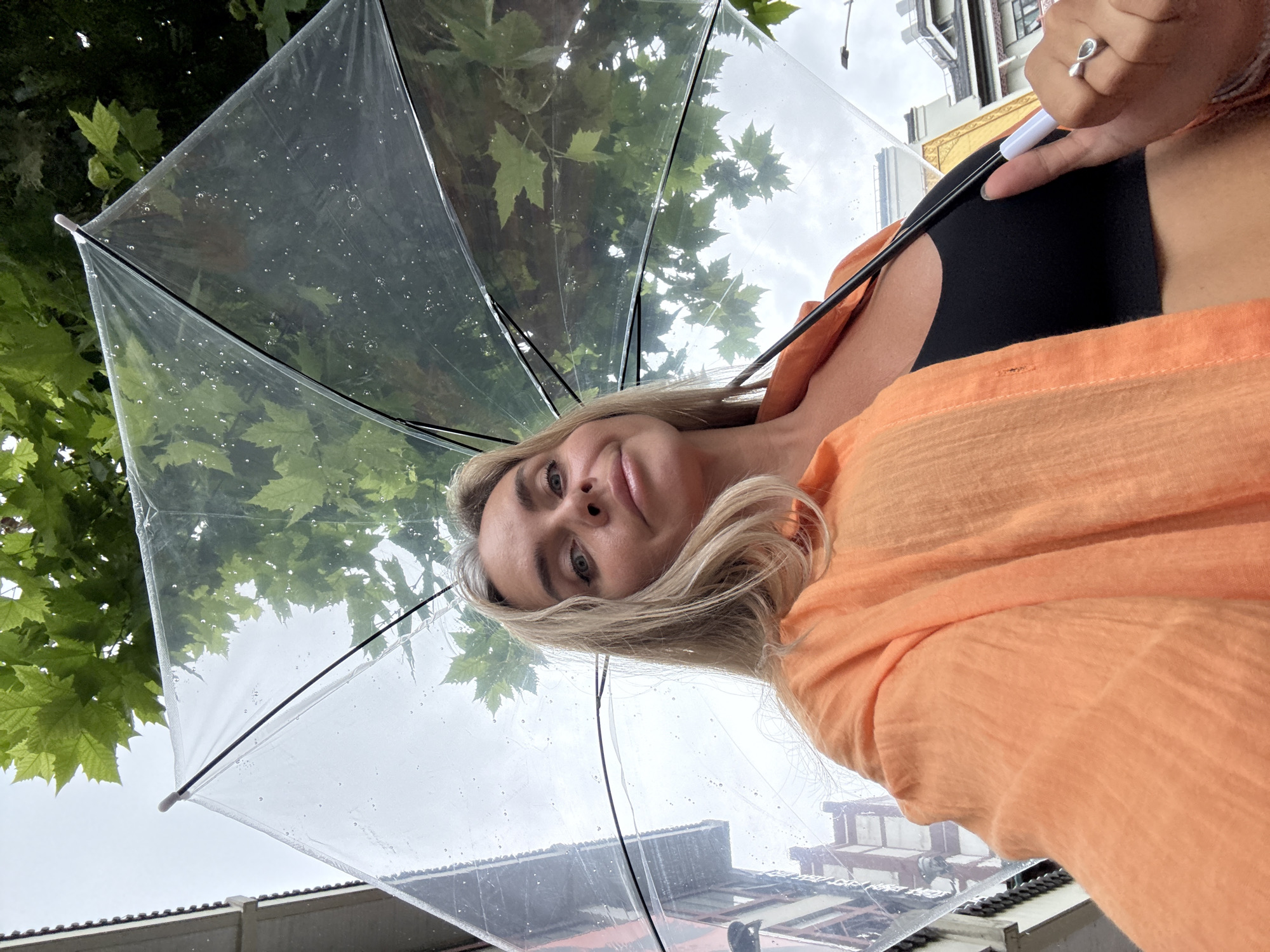 A medium shot photo shows a woman from the shoulders up, holding a transparent umbrella open above her head. She has blonde hair and is looking at the camera. She is wearing an orange shirt over a black top. The umbrella is speckled with raindrops, and green leaves are visible through it from trees overhead. The background shows buildings and a cloudy sky. The image was taken outdoors.