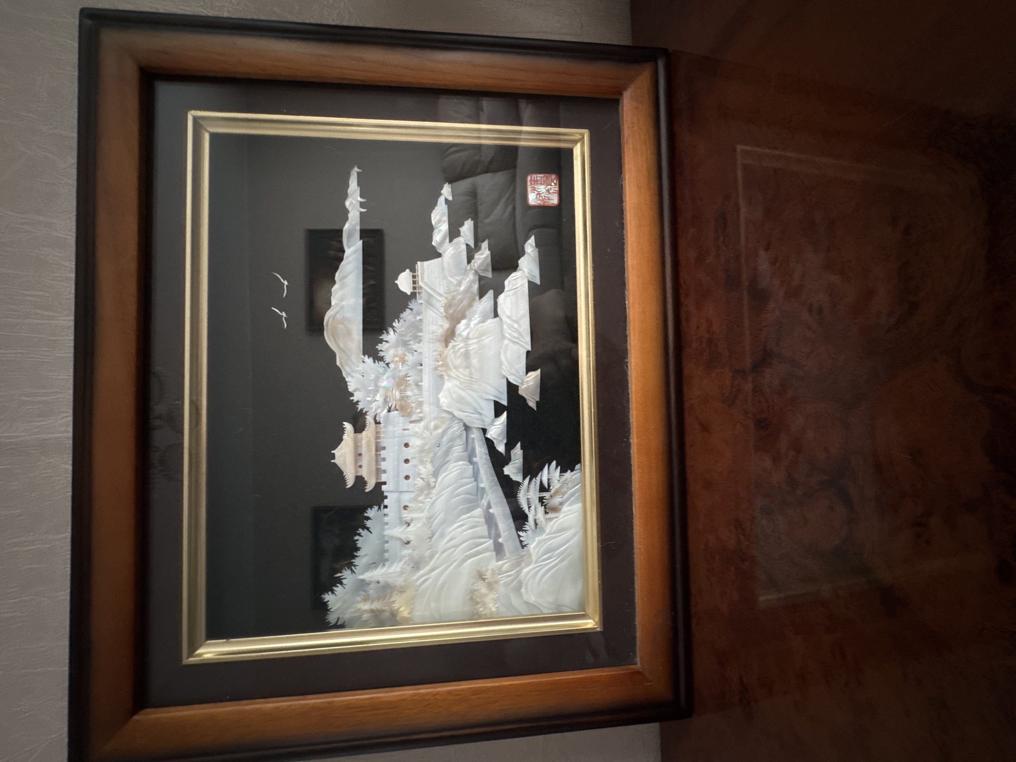 The image shows a framed artwork, likely depicting a landscape scene with a temple, a wall, and mountains, created using what appears to be mother-of-pearl or a similar iridescent material. The artwork is housed in a dark brown frame with a lighter wood trim. The background of the artwork is black, and the relief artwork is primarily white and iridescent. There are two small birds depicted in flight in the upper right portion of the artwork. A red stamp or seal is visible in the lower right corner of the artwork. The framed artwork is placed on a polished wooden surface, possibly a table or desk, which reflects the artwork and the surrounding environment. The surface has a rich, dark brown color with a visible wood grain pattern. The overall setting appears to be indoors, with a textured wall visible in the background above the frame. The image does not contain any information that would indicate its location in the city of Novosibirsk, Russia.