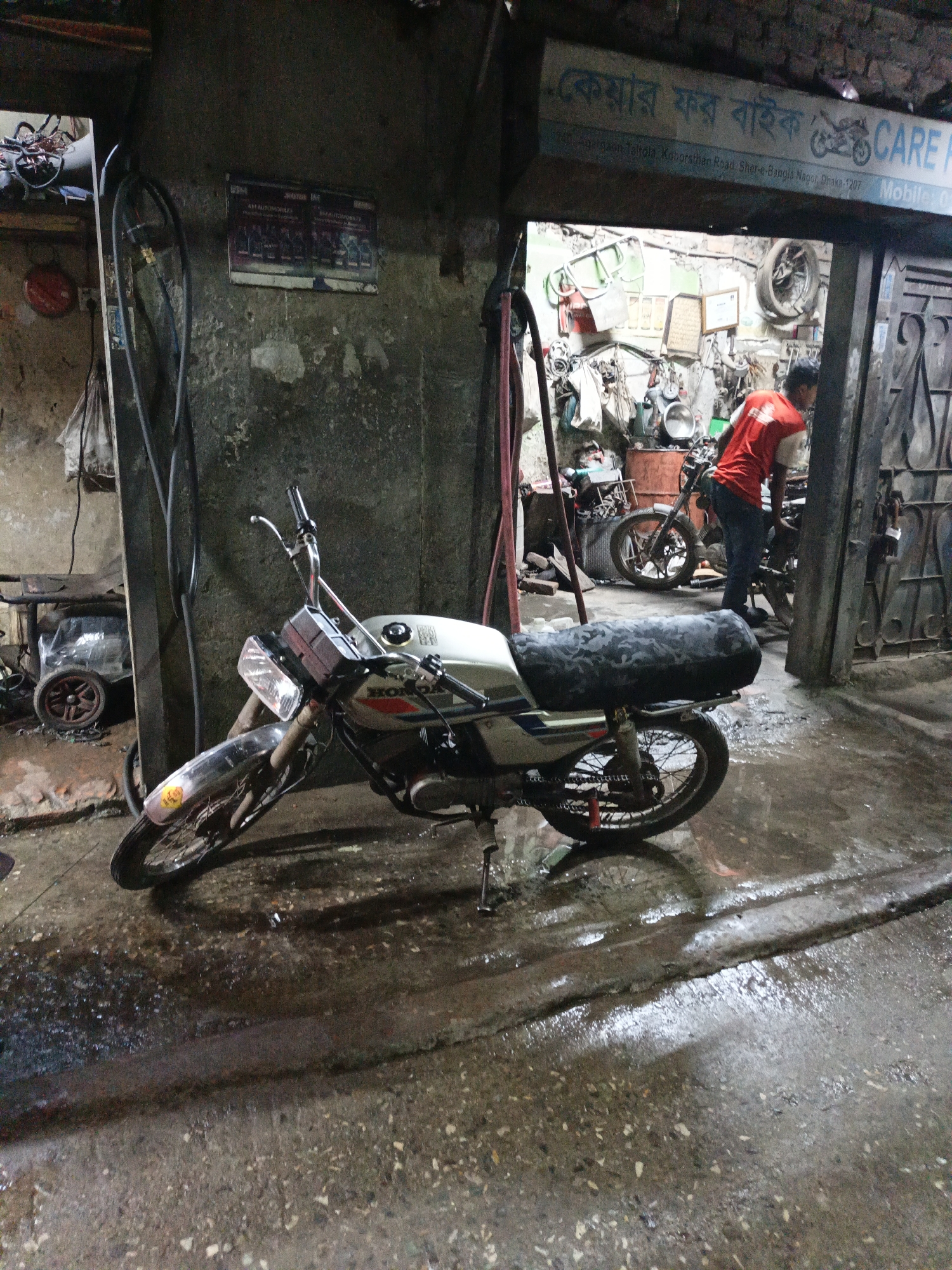 A motorcycle repair shop in Dhaka, Bangladesh is viewed from outside the open entrance. A white motorcycle is parked in front of the entrance, its front tire turned sideways. The ground is wet. Inside the shop, a man wearing a red shirt and blue jeans works on a motorcycle. Other motorcycles and parts are visible. A sign above the entrance reads "কেয়ার ফর বাইক CARE." The sign also lists an address: 240 Agargaon Taltola, Koborsthan Road, Sher-e-Bangla Nagor, Dhaka-1207. Another sign is affixed to the concrete wall to the left of the entrance. An air compressor on wheels is near the left wall.