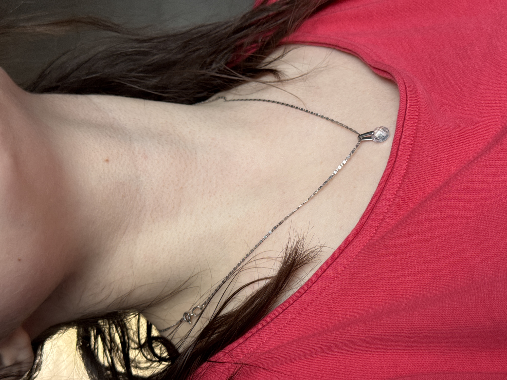 A close-up, angled shot captures the upper chest and neck area of an individual, seemingly in Izhevsk, Russia. The person, whose face is not visible, is wearing a red, possibly cotton, t-shirt or garment. A delicate, silver-toned chain necklace with a prominent, clear, faceted crystal pendant rests on their collarbone area. The pendant, spherical or teardrop-shaped, catches the light. Dark brown, long hair cascades around the neck and chest, with some strands lying across the skin and garment. The skin shows natural texture and a small mole is visible on the neck. The image is taken from an angle, with the person's head area positioned towards the left side of the frame, and their body extending towards the right. The background behind the hair is dark and indistinct. There is no visible text.