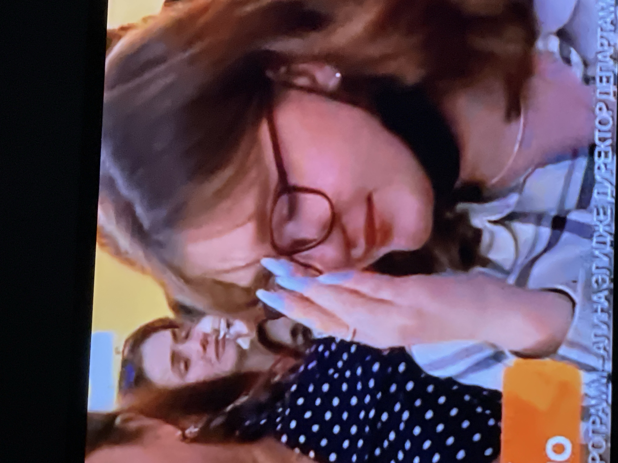 The image shows a close-up of a young woman with light brown hair and glasses, wearing a striped shirt and a black face mask around her neck. She is touching her face with her right hand, which has long, light blue fingernails. She appears to be in a public gathering, as other people are visible in the background. The setting appears to be indoors or possibly a covered outdoor area, with a yellow background behind the subjects. There is text at the bottom of the image which is partially visible and appears to be in Russian. The specific location within Moscow, Russia, cannot be determined from the image alone.