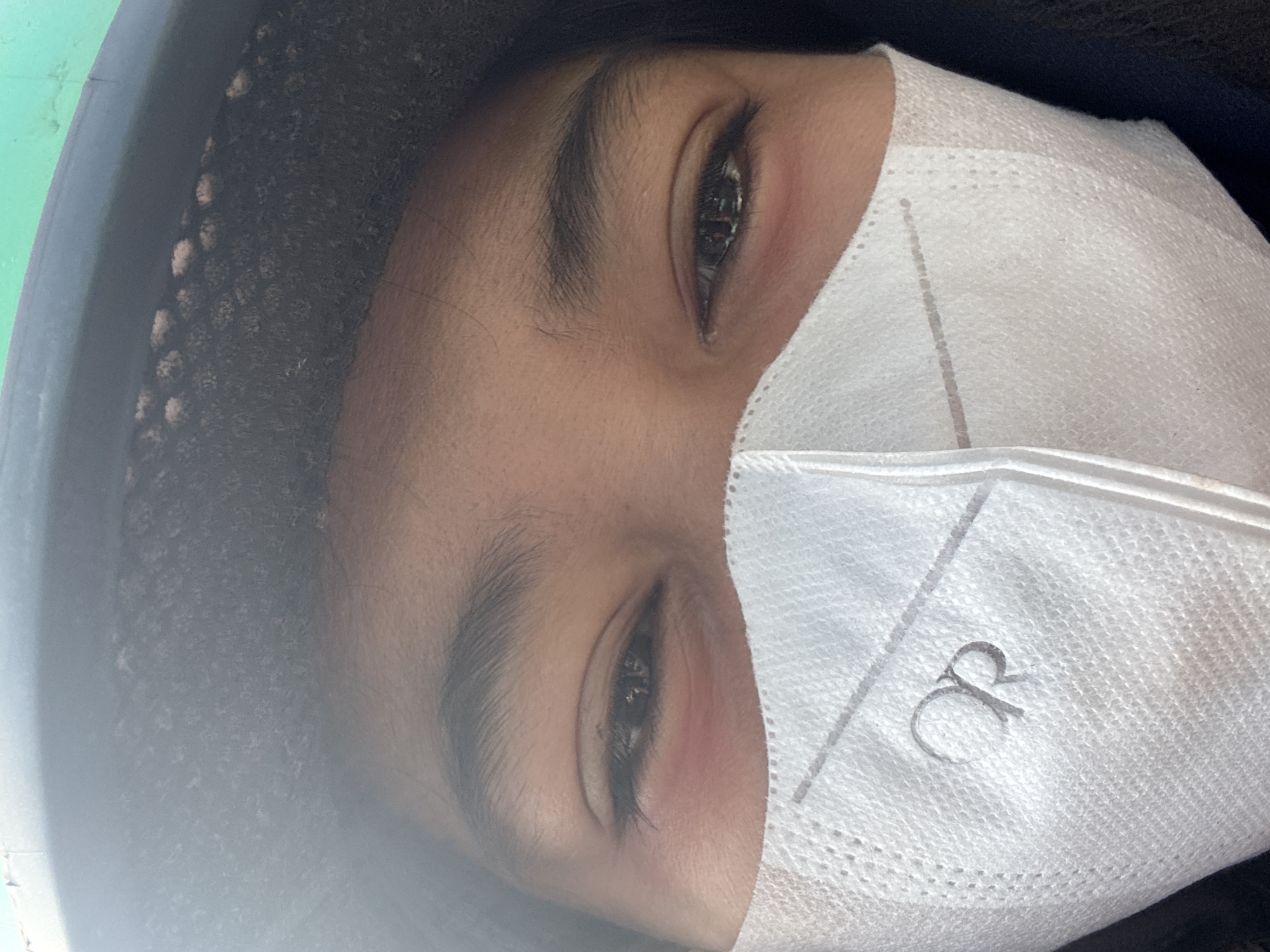 A close-up, eye-level, indoor shot captures a person wearing a white face mask that partially obscures their face. The mask features a grey logo resembling the letters "OR." Their eyes are visible above the mask, with dark eyeliner applied. The person is wearing a dark helmet, and a portion of a green object is visible at the top of the frame, suggesting they may be on a motorcycle. The background is blurred and appears to be an indoor setting. The image quality is adequate, with good lighting and focus on the subject's eyes and mask.