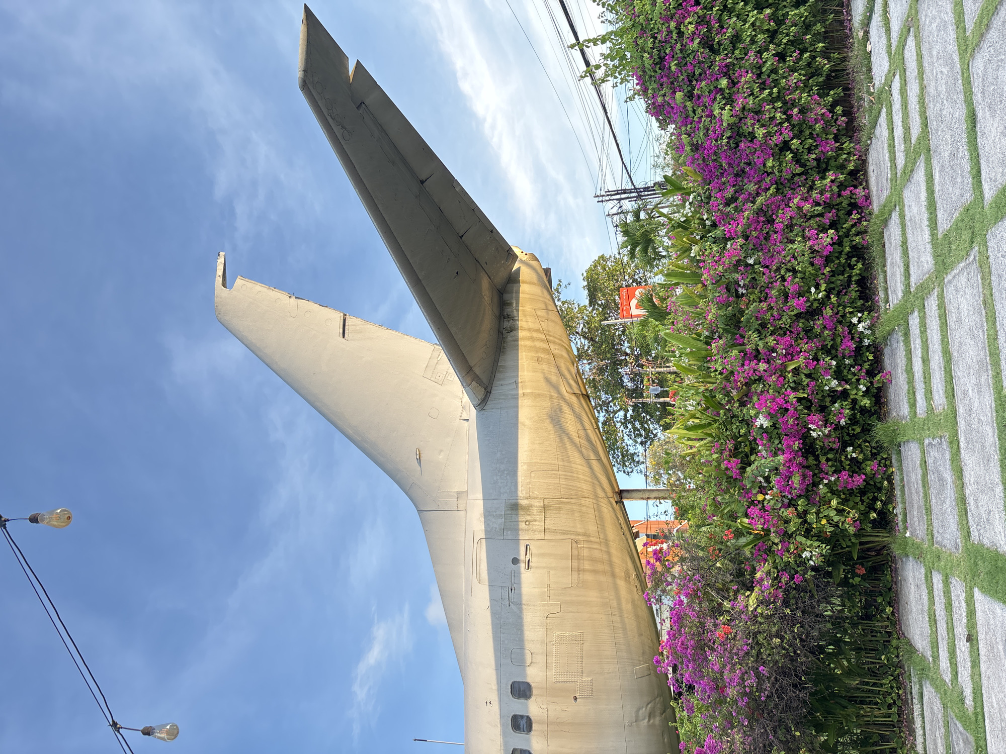The image captures the tail section of an aircraft juxtaposed with lush vegetation in an urban setting. The tail, painted in a light color, is prominently featured against a backdrop of a bright blue sky with wispy clouds. The tail is partly covered in bougainvillea flowers, and next to the tail is a pathway made of light stone and grass, indicative of a garden or landscaped area.
The location can be identified as the former site of the “Gate 88 Airplane Park” in Kuta, Indonesia. It prominently features a Boeing 737 tail. The park has since closed, and the aircraft section has been converted into a roadside attraction or potentially part of a new establishment. There is no evidence of any people in the frame.
