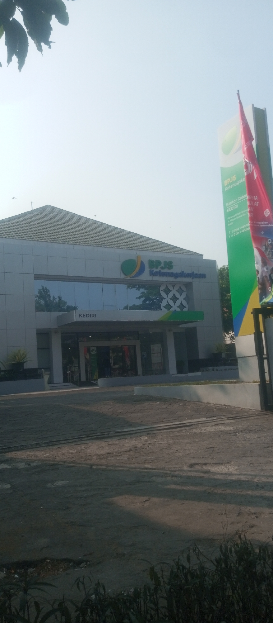The image shows the exterior of a BPJS Ketenagakerjaan office in Kediri, Indonesia. The building is modern with a light-colored facade and a dark-tiled roof. Large glass windows reflect the surroundings. Above the entrance, the word "KEDIRI" is prominently displayed. To the right of the building, a tall sign displays the BPJS Ketenagakerjaan logo and information about the office, including its address and contact details. A red flag is attached to the signpost. The foreground shows a paved area, possibly a parking lot or walkway, with some vegetation. The sky is bright and clear.