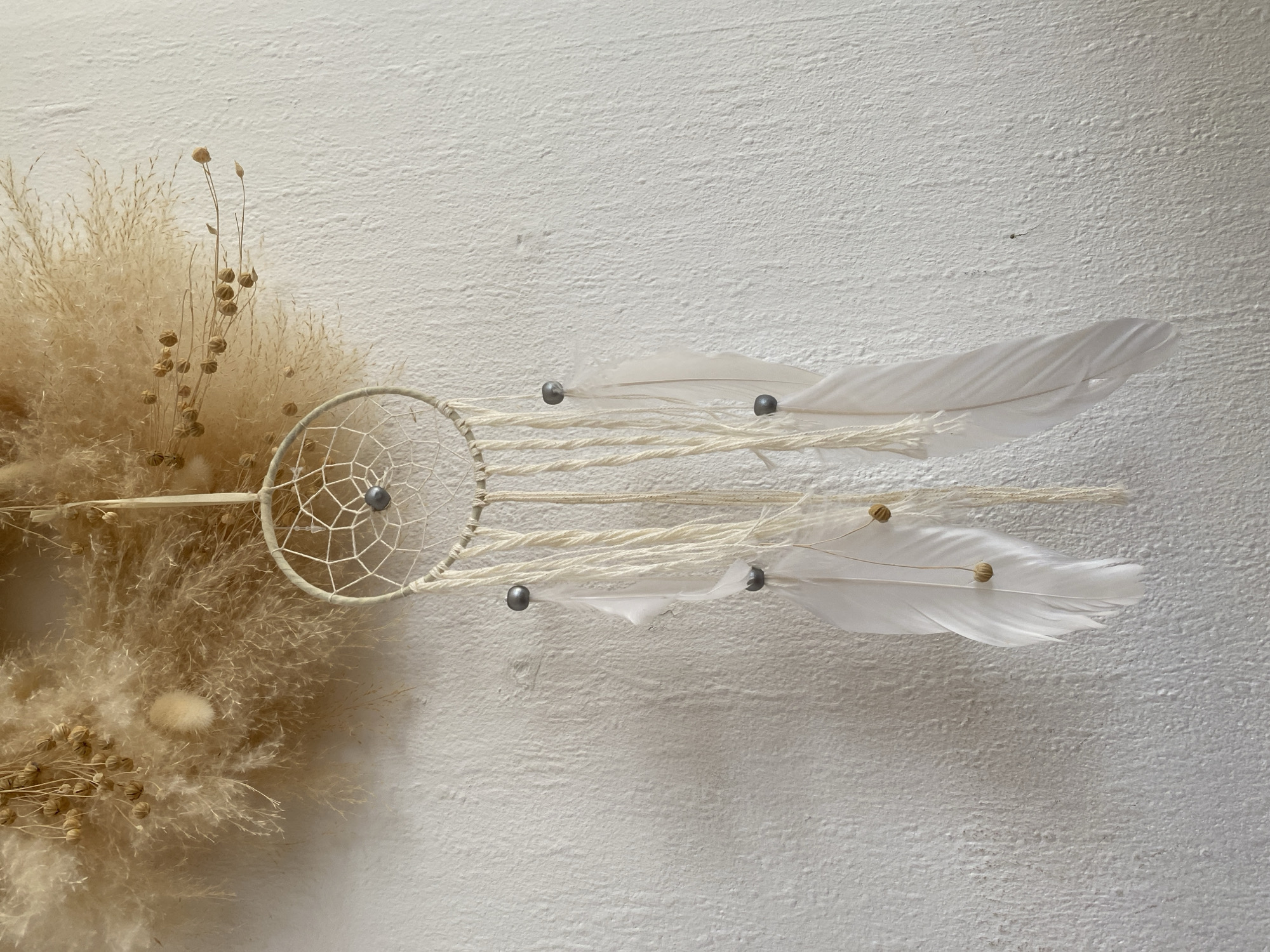 A dreamcatcher is hanging on a textured white wall in Bad Sachsa, Germany. The dreamcatcher is circular with a white string web inside, and decorated with white feathers and small dark beads. Above the dreamcatcher, there is a decorative arrangement of dried grasses and seed pods. The lighting suggests daytime. There are no people or animals visible in the image.