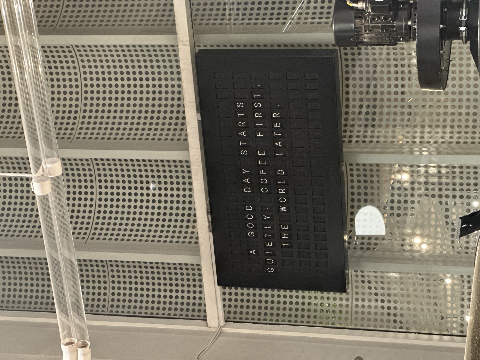 The image depicts a close-up view of a ceiling, likely inside a building, with a black, rectangular sign displaying text. The sign reads: "A GOOD DAY STARTS QUIETLY: COFFEE FIRST, THE WORLD LATER." To the left of the sign are two vertical, transparent tubes secured with white bands, possibly part of a lighting fixture. The ceiling itself is made of a perforated material with small circular holes. In the upper right corner, parts of an industrial-looking motor or machine are visible. The image provides no evidence to confirm its location in Yerevan, Armenia.