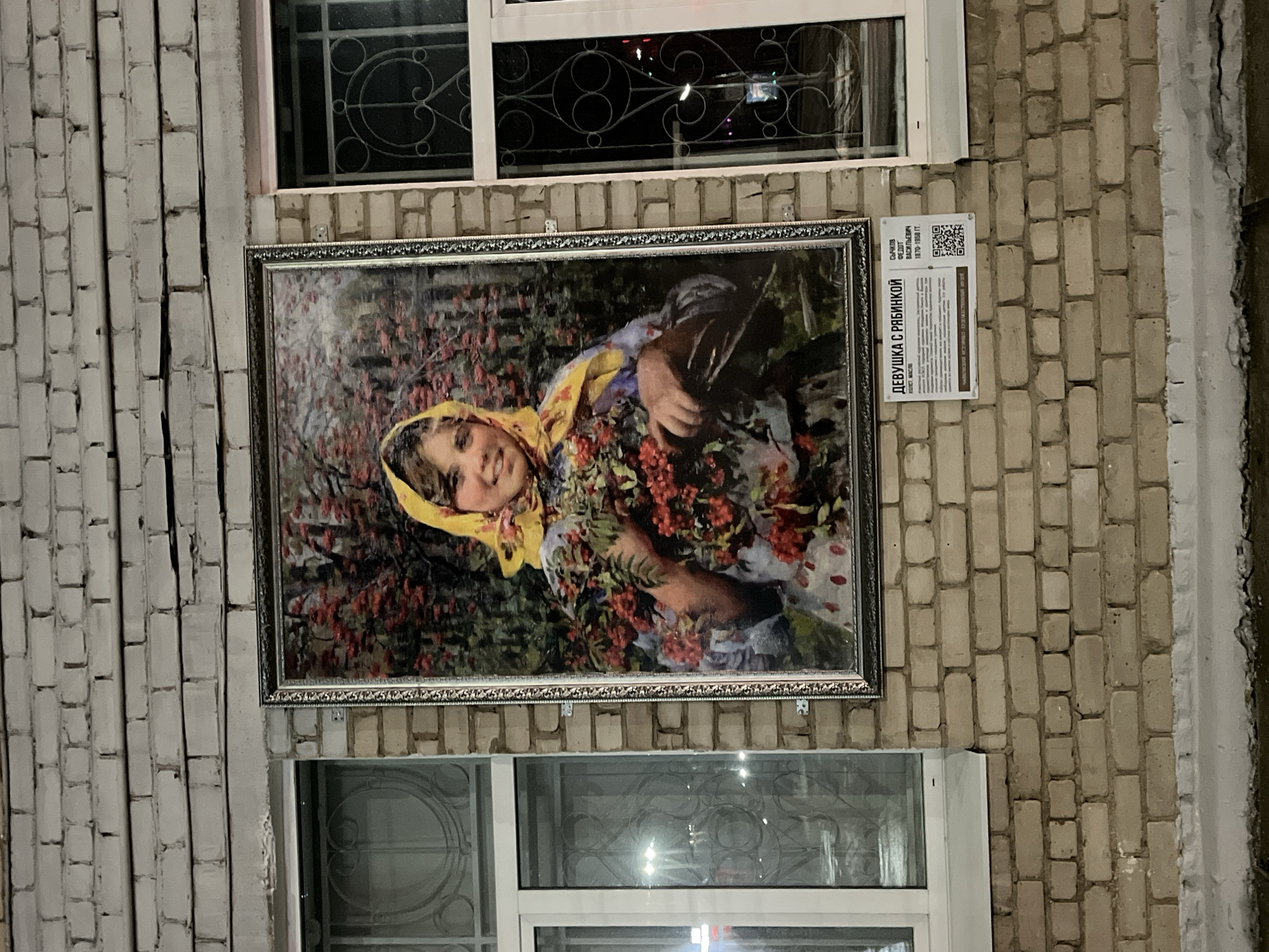 The image shows a framed picture of a girl with a yellow headscarf holding red berries. The painting is mounted on a brick wall between two windows. To the right of the picture, there's a sign with text and a QR code. The location is in an unidentified part of the city of Unknown, Unknown.