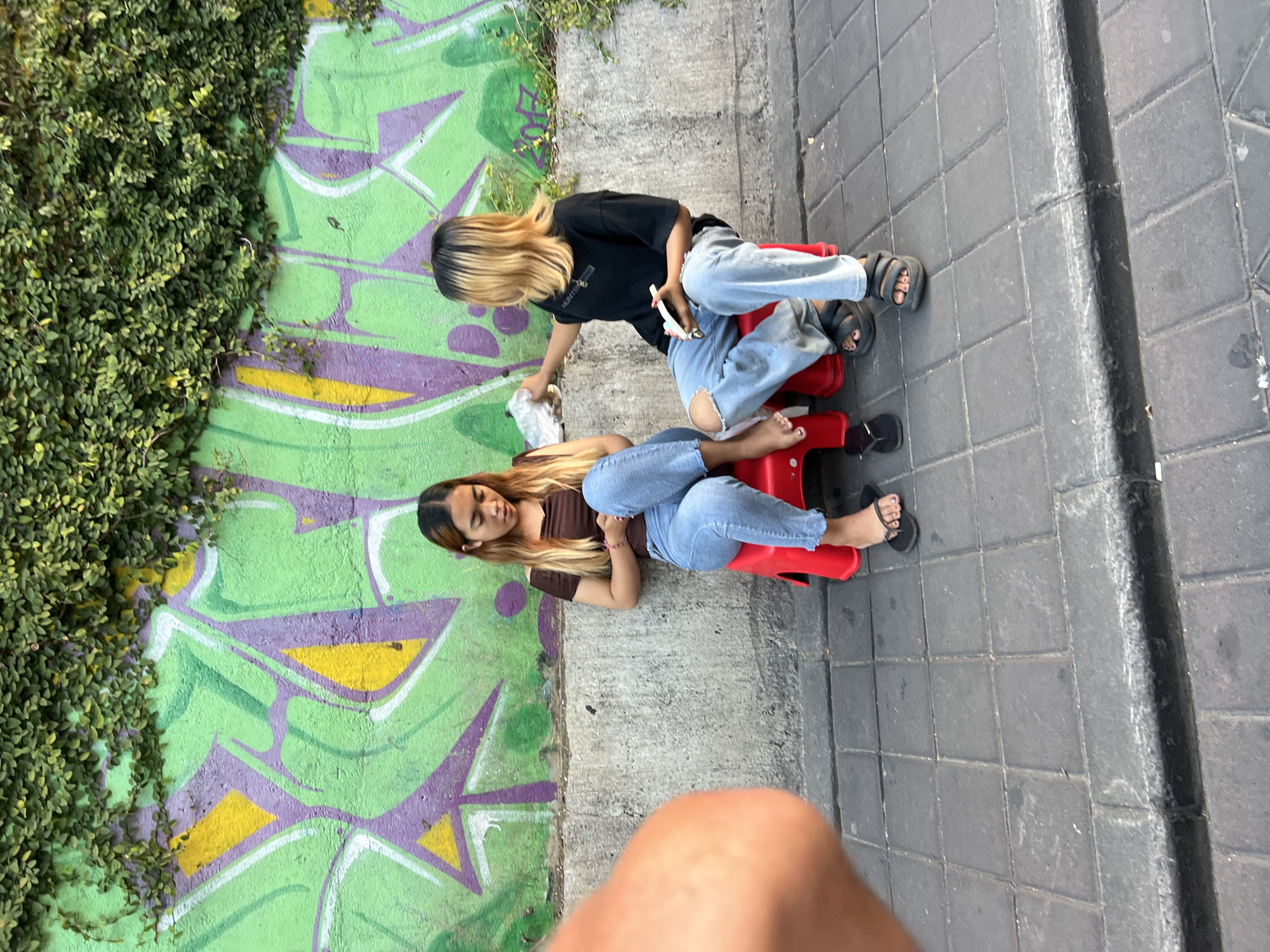 Two young women are sitting on red plastic stools in front of a concrete wall covered in green ivy and graffiti art in Kuta, Indonesia. The woman on the left, wearing a brown off-the-shoulder top and blue jeans, is looking down at her lap. The woman on the right, wearing a black t-shirt and ripped blue jeans, is holding a small white object in her right hand and looking towards the left. The ground is paved with gray square tiles. The scene appears to be a street or sidewalk.