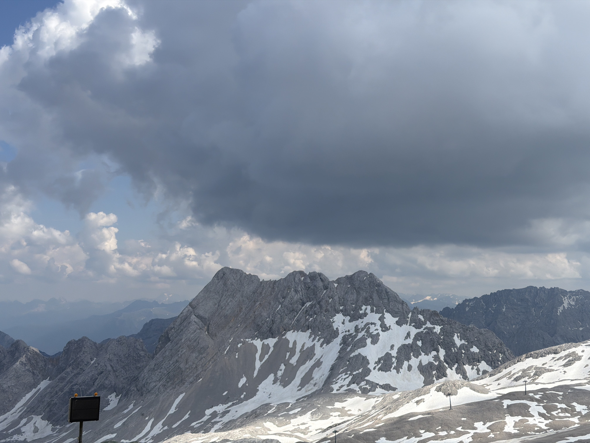 This media file depicts a panoramic view of a rugged, snow-dusted mountain range under a dramatic sky. The foreground features rocky slopes partially covered in patches of snow, suggesting a high-altitude environment. Two prominent poles with lights are visible in the lower part of the frame, likely indicating a trail marker or part of a ski lift system.
In the middle ground, a majestic, jagged mountain peak dominates the scene. Its grey rocky surfaces are interspersed with white snow, creating a stark contrast. Further in the background, successive layers of mountains recede into a hazy blue distance, emphasizing the vastness of the landscape.
The sky is a significant element, filled with a mix of dark, imposing storm clouds and lighter, fluffy cumulus clouds scattered against a pale blue backdrop. This suggests a dynamic weather system, possibly indicating an approaching storm or clearing after one. The lighting is subdued, consistent with an overcast day or the late afternoon.
The location is identified as Garmisch-Partenkirchen, Germany, a region known for its alpine scenery and outdoor activities. The presence of snow on the mountains, even in what appears to be warmer months, points to the high elevation typical of this area. No people are visible in the image. The overall mood is one of awe-inspiring natural grandeur, tinged with the dramatic intensity of the weather. There is no visible text in the image.