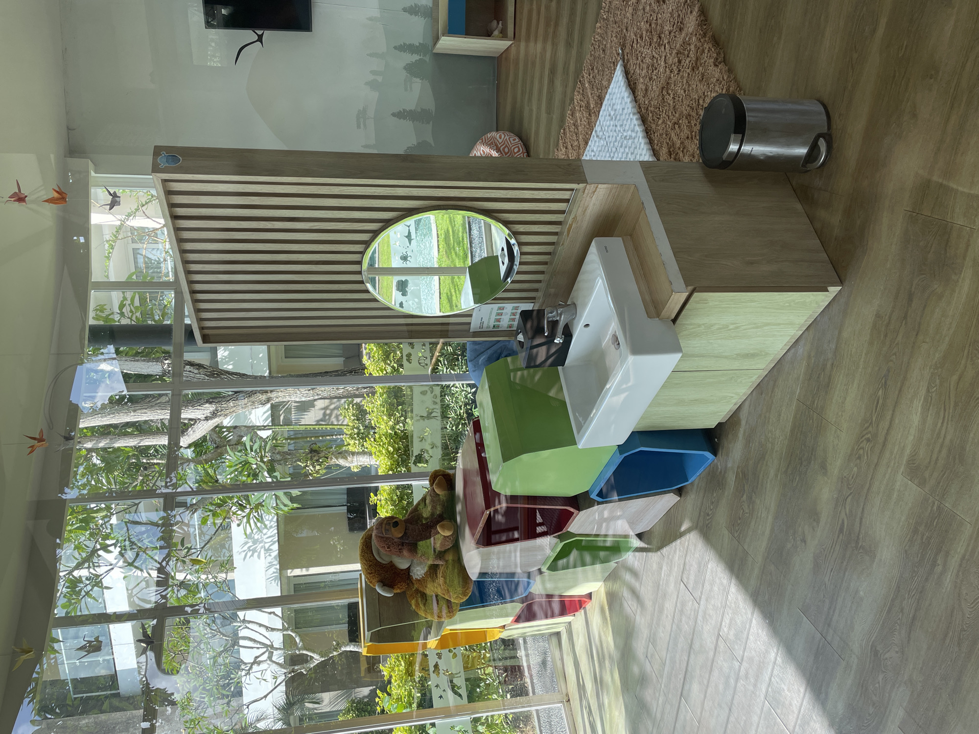 The scene appears to be an indoor setting, likely a bathroom or washroom in a commercial or public facility. The foreground is dominated by a modern sink and vanity unit with a wooden countertop and striped wooden back panel, reflecting an oval mirror. The sink is white with a visible tap. Below the sink, there are colorful, geometric-shaped containers stacked together. A stuffed animal is perched on top of the stack, next to a large window.
The background shows a natural landscape mural on the wall, featuring trees and mountains. There's also a tall window behind the fixtures that reflects the outside view with lush greenery and architectural structures.
The environment suggests a child-friendly design, possibly in a daycare, kindergarten, or a similar venue with modern and artistic elements. The location appears to be in an area with abundant greenery, and there are decorative origami birds near the window. No people are visible in the scene.