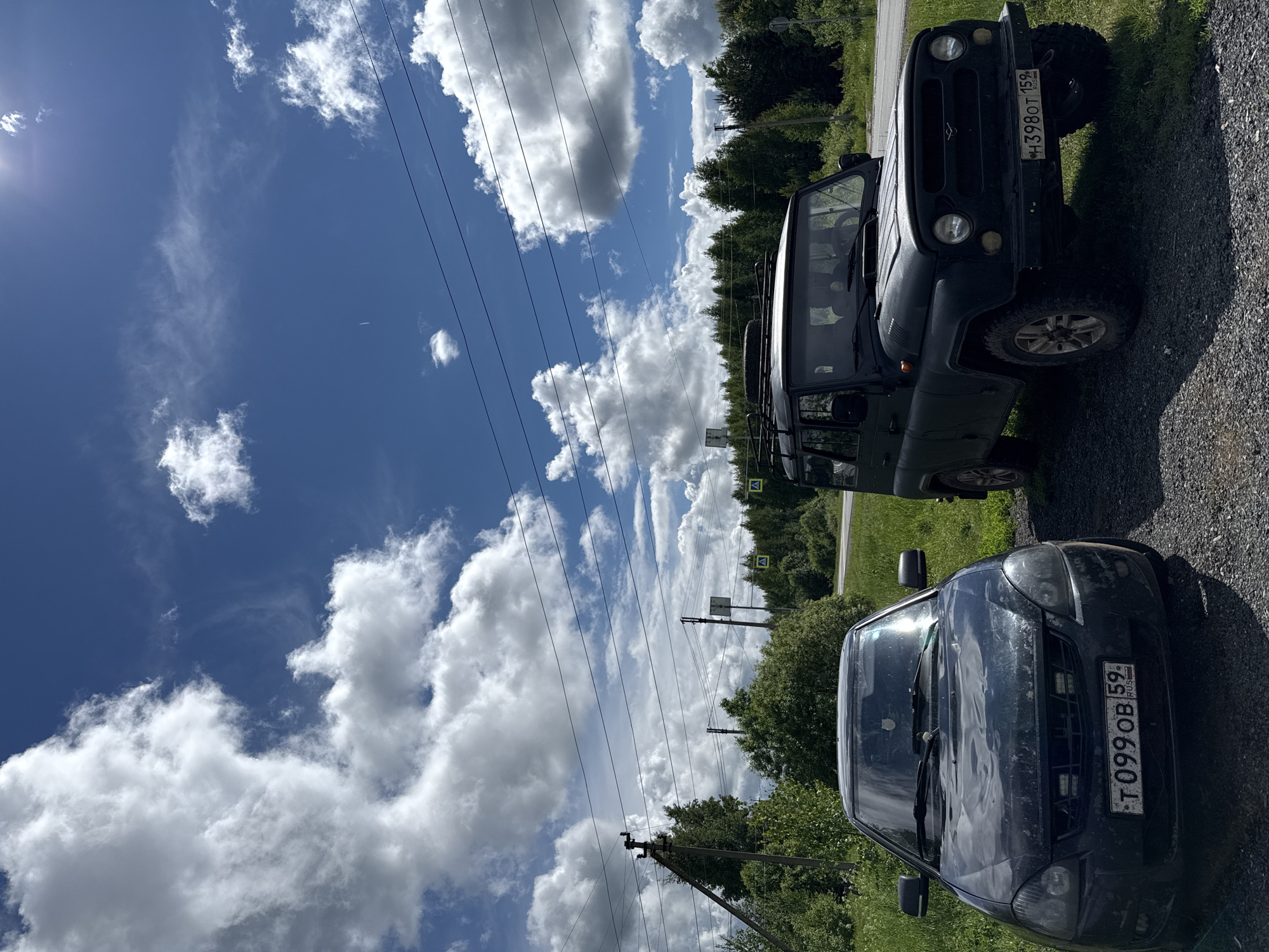 The image depicts a bright, sunny daytime scene in what appears to be a rural or semi-rural setting, likely alongside a road. The sky is a vivid blue, adorned with numerous fluffy white cumulus clouds. Power lines stretch across the upper and middle sections of the frame, supported by visible utility poles.
Two dark-colored vehicles are parked on a gravel shoulder. In the foreground, positioned to the right, is a dark sedan, possibly a Lada. Its front shows some dust or dirt, and its license plate reads "T099OB 59 RUS." Behind it and to the left is a dark green, rugged SUV, identifiable as a UAZ, with its front facing towards the viewer. The SUV's license plate displays "H398OT 159 RUS." Both license plates feature the "RUS" country identifier, and the "59" and "159" regional codes are consistent with Perm Krai in Russia, aligning with the Zhigalki location context.
Lush green trees and dense vegetation form a backdrop along the roadside. A distinct sun flare is visible in the upper left quadrant of the sky. Several yellow diamond-shaped road signs, possibly warning signs, are partially visible among the trees in the mid-ground. The scene is devoid of people, conveying a quiet, natural environment on a clear day.