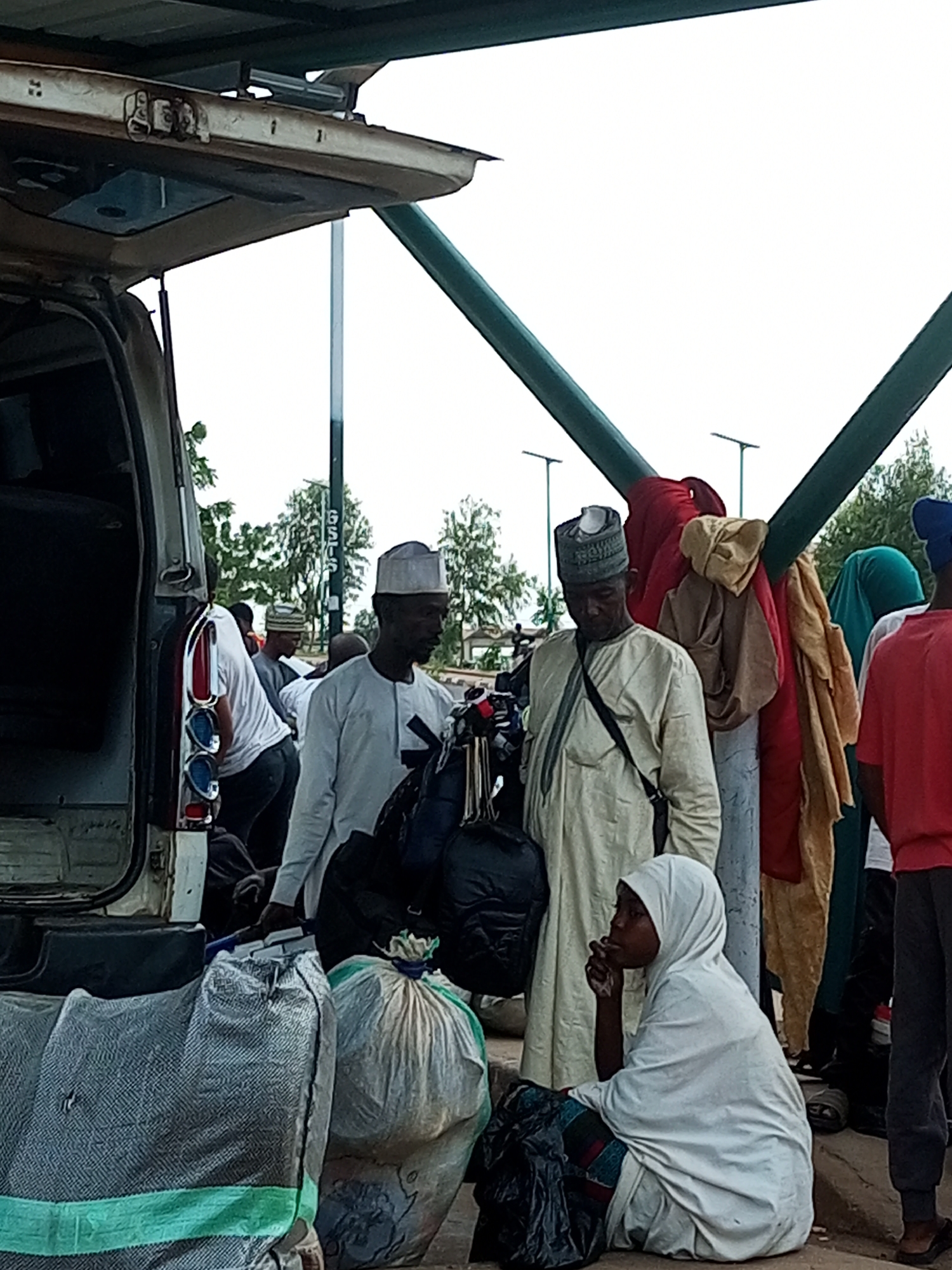In Gombe, Nigeria, a group of people are gathered near an open vehicle, possibly preparing for travel or unloading belongings. The scene appears to be outdoors, under a green-colored awning or shelter, with a street or road visible in the background, along with trees and streetlights. The people are dressed in traditional attire, with several men wearing caps and robes, and one woman in a hijab. They are surrounded by various bags and luggage, suggesting they are in transit. Some individuals are standing and conversing, while others are seated. The overall atmosphere suggests a moment of waiting or transition.