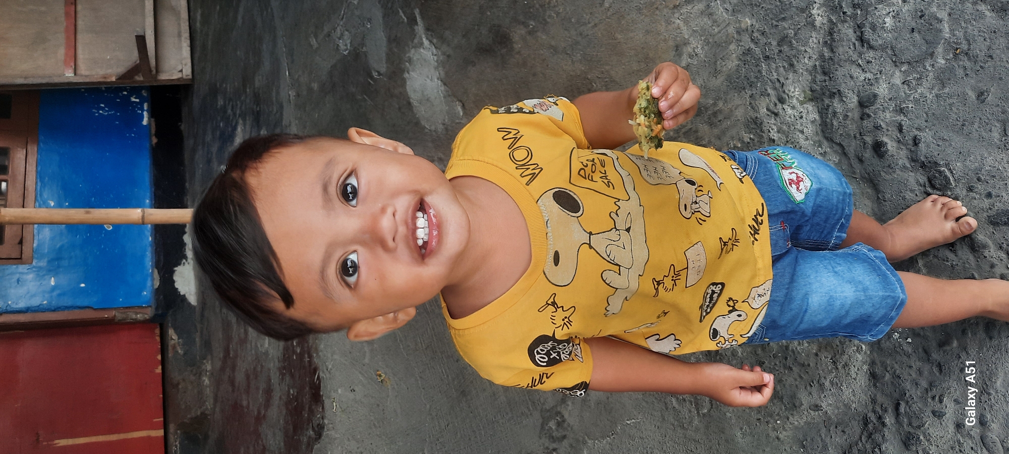 A young child, likely a boy based on clothing, stands barefoot on a textured gray concrete surface. He is wearing a bright yellow t-shirt adorned with a repeating pattern of Snoopy characters and text, alongside blue denim shorts with a patch on the right leg. The child is holding a small, green, leafy snack in his right hand and is looking up and smiling at the camera, revealing his teeth. In the background, a blue wall with some peeling paint is visible, as well as a red door or wall section and a wooden pole. The overall setting appears to be outdoors in a rustic environment, potentially a residential area in Selopuro, Indonesia, given the context. The lighting suggests it is daytime.