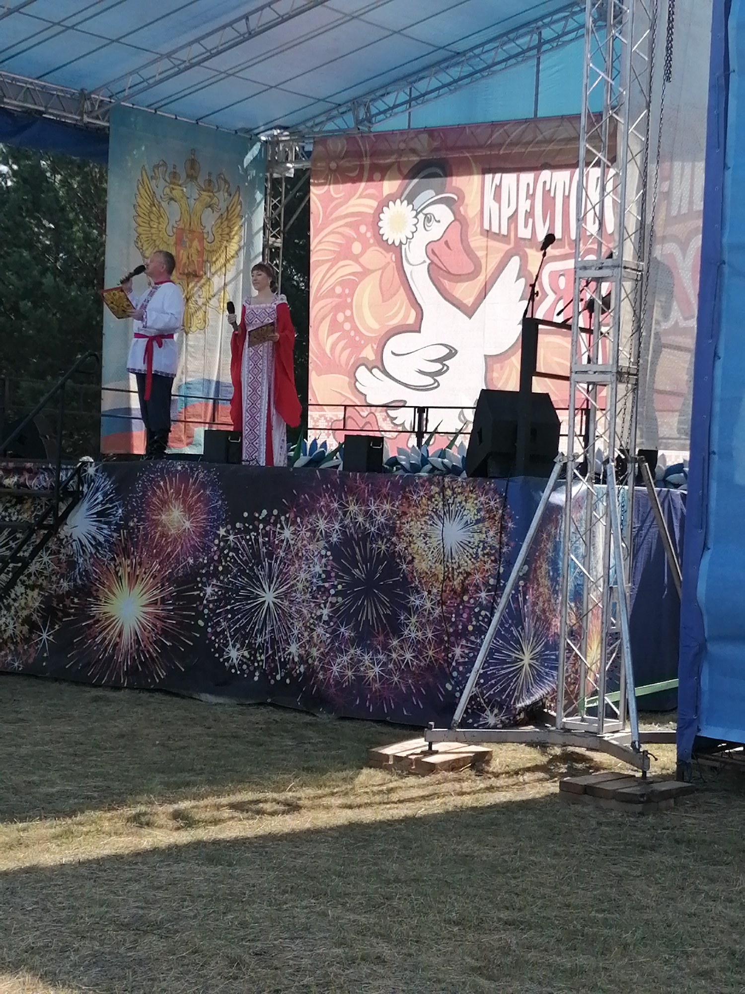 On an outdoor stage, two individuals in traditional Russian attire are performing. On the left, a man in a white embroidered shirt with a red sash holds a microphone and a book. To his right, a woman in a red and white patterned dress also holds a microphone and a book. They are standing in front of a backdrop featuring a large image of a white goose wearing a hat with a flower and the Cyrillic word "КРЕСТОМ" (KRESTOM). To the left of the goose image is a banner displaying a golden double-headed eagle. The stage is decorated with a backdrop of colorful fireworks. The stage structure itself is a metal truss system. The ground in front of the stage is covered in dry grass, with shadows cast by the stage and its supports. The scene appears to be at an event in Shadrinsk, Russia.