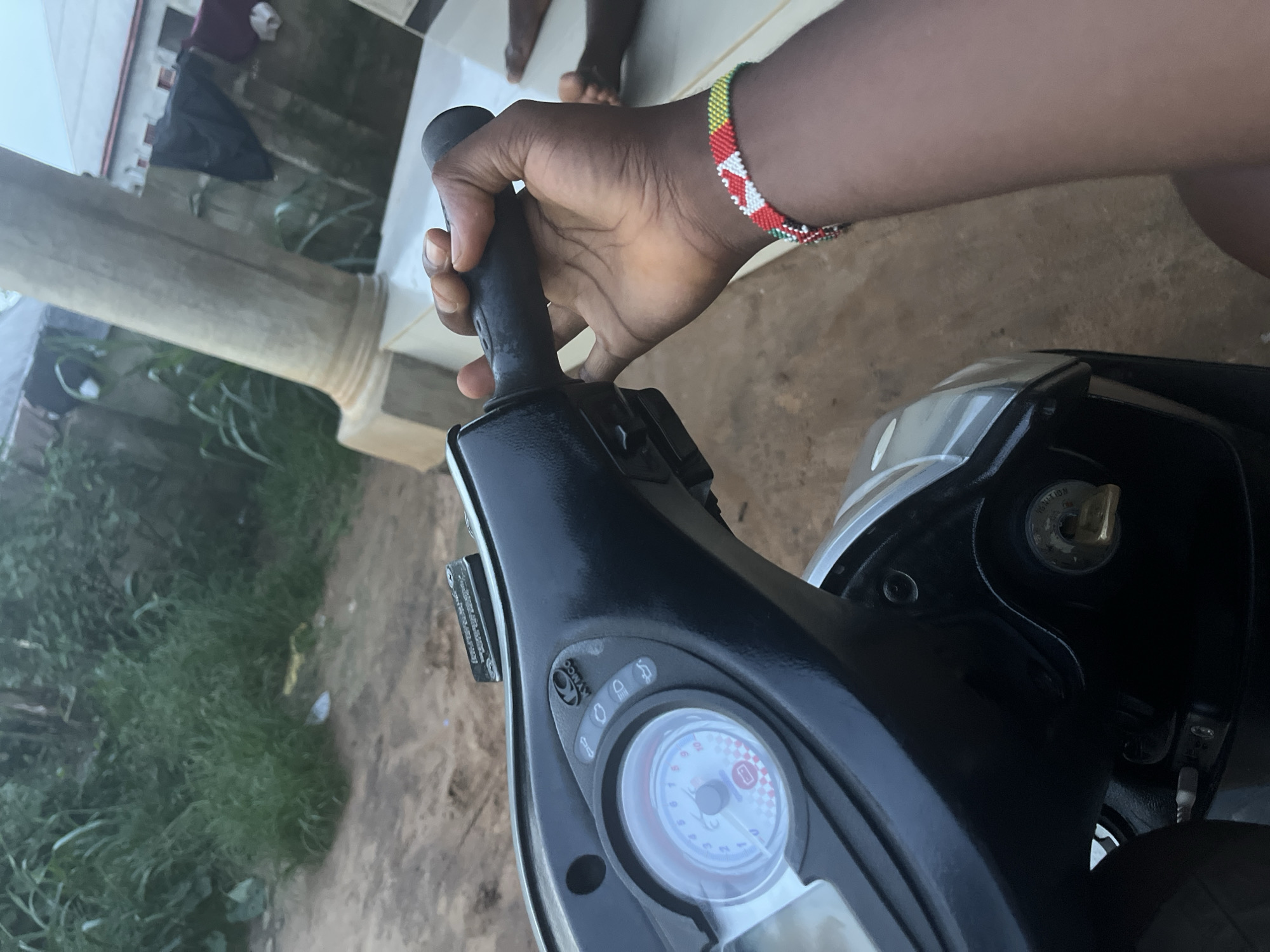The image presents a close-up, first-person perspective of a person's dark-skinned arm and hand gripping the right handlebar of a scooter. A vibrantly colored beaded bracelet, featuring red, white, green, and yellow patterns, is worn on the wrist. The scooter's dashboard is visible, displaying a speed dial (partially obscured), indicator lights, and the "KYMCO" brand logo. An ignition key is inserted into the keyhole, which is labeled "IGNITION."
The scene is set outdoors in what appears to be a residential or semi-urban area in Ikot Ekpene, Nigeria. To the left, a sturdy light-colored concrete pillar suggests the presence of a building or veranda. Beyond this, a lush expanse of green grass and wild foliage fills the background. The immediate ground under the scooter is an unpaved, earthy surface, transitioning to a lighter, possibly tiled or concrete area closer to the distant building. Parts of two other people's bare feet are visible in the background, implying additional individuals are present near the structure. The lighting is soft and diffused, suggesting either an overcast sky or the early morning/late afternoon hours. No strong shadows are present. A dark piece of fabric is also discernible near the building in the distance.