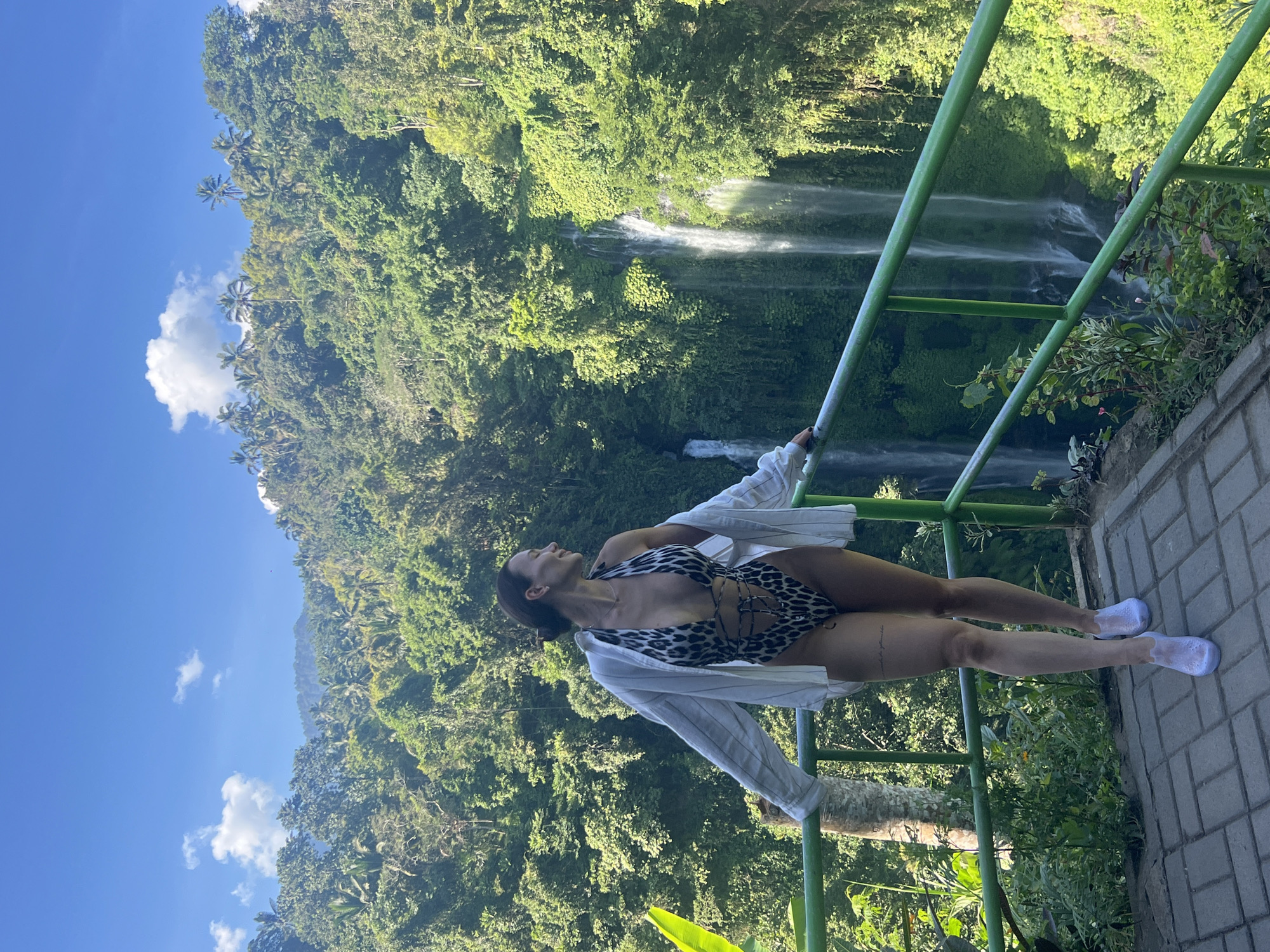 A woman stands on a paved overlook, leaning on a green railing, facing a lush green jungle with multiple waterfalls. She is wearing a leopard-print one-piece swimsuit and an unbuttoned white linen shirt. Her hair is pulled back, and she is looking to her right with a slight smile. The background features dense tropical vegetation and several cascading waterfalls. The overlook has a patterned brick floor and is bordered by a railing and surrounding greenery. The sky is blue with scattered white clouds. The location appears to be a natural scenic viewpoint, likely a tourist attraction in Sawan, Indonesia, known for its natural beauty and waterfalls.