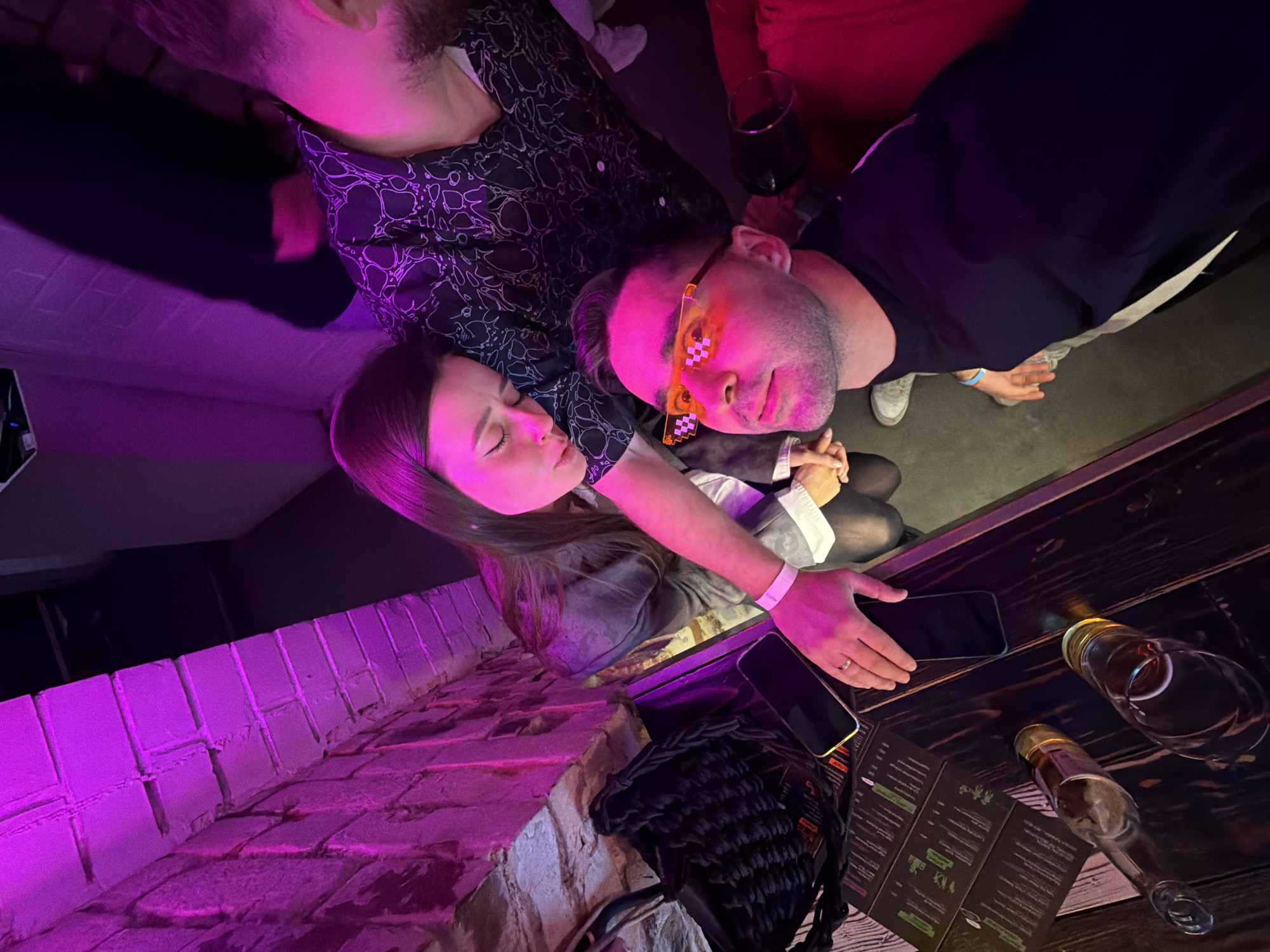 The image captures an indoor, dimly lit scene, likely a bar or club in Krasnoyarsk, Russia, illuminated by vibrant purple and pink neon lights. The photograph is taken from a slightly elevated, tilted perspective, looking down onto the subjects and a bar counter.
Three individuals are prominent. On the left, the top of a man's head and his beard are partially visible. Next to him, a woman with long dark hair rests her head, eyes closed, on a second man's shoulder. She wears a light-colored top under a dark jacket and has a white wristband on her left arm. The central man, wearing a dark shirt, is seen looking towards the camera, sporting unique orange pixelated glasses that reflect the ambient light. His expression appears neutral, possibly with a slight smirk. A fourth person's arm, adorned with a blue wristband, is visible near the central man's right shoulder.
The activity appears to be socializing and relaxing. The woman seems to be comfortably leaning, perhaps tired, while the man with glasses engages with the camera.
In the foreground and midground, a dark wooden bar counter holds several items: two smartphones, a dark woven bag or item, a folded menu (with text unreadable), an empty clear glass, and two clear bottles (one empty, one possibly containing an amber liquid). A red wine glass, filled, is situated near the central man. The background features a brick wall, intensely washed in purple light, and beyond that, hints of red upholstery, likely seating. The artificial lighting indicates a nighttime setting.