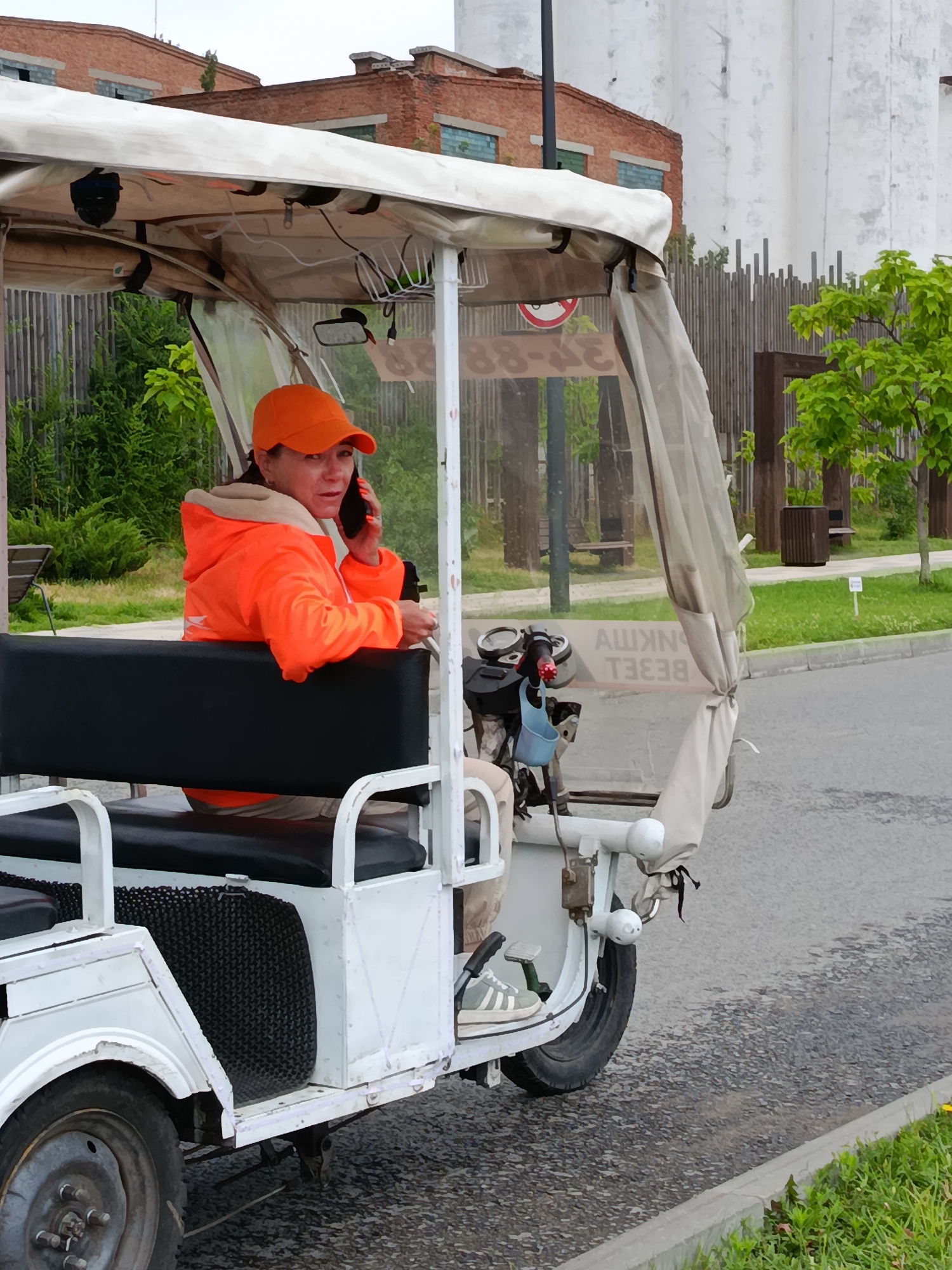 A woman wearing an orange baseball cap and an orange jacket is sitting in the driver's seat of a white, three-wheeled vehicle, talking on a black cell phone. The vehicle has a white canopy overhead and a black seat. The woman is looking forward and to her left. The vehicle is parked on an asphalt road next to a curb with green grass. In the background, there are buildings, trees, and a wooden fence. The overall lighting suggests an overcast day.