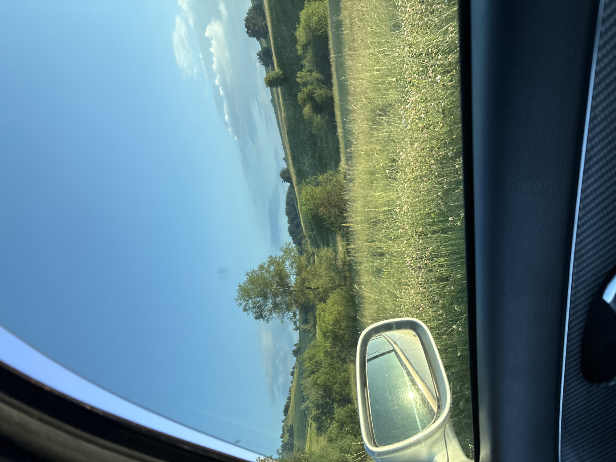 The image captures a view from inside a vehicle, looking outwards towards a sunny, rural landscape in Koprivshtitsa, Bulgaria. The perspective suggests the vehicle is moving, as indicated by the side mirror reflecting the road and the blurred greenery.
The scene is dominated by a vast expanse of clear blue sky with scattered white clouds, suggesting good weather and daylight, likely mid-morning or afternoon due to the bright sunlight. Below the sky, a rolling green landscape unfolds, characterized by fields of tall grass and scattered trees. The grass appears to be dry and light brown in places, hinting at the dry summer season. The fields are not uniform, with some areas appearing more densely vegetated than others, creating a textured visual.
A prominent object in the foreground is the vehicle's side mirror, positioned on the right side of the frame. It is a standard car mirror with a silver casing, reflecting the sunlit road and the surrounding green environment. The interior of the car is also visible, with a black dashboard and a textured trim, possibly carbon fiber, on the right side of the frame.
The overall mood is one of tranquil travel and appreciation of the natural scenery. The image evokes a sense of a pleasant journey through the Bulgarian countryside, highlighting the beauty of the natural landscape under a bright sky. There is no visible text in the image.