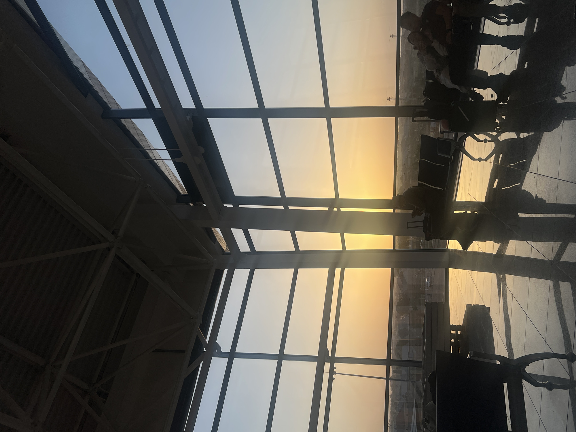 The image displays the interior of an airport terminal in Shymkent, Kazakhstan, during sunrise or sunset, characterized by strong backlighting. Golden sunlight streams through a large, multi-pane window wall that occupies the entire right side of the frame, casting long shadows and bright reflections on the polished, light-colored floor. The ceiling reveals exposed white structural trusses. Several dark-colored public seating benches are occupied by individuals, rendered in silhouette against the bright light. In the mid-right, a person sits alone, with a black wheeled suitcase next to them. Further right, a couple is seated closely together, with a backpack visible near their feet. A partially obscured individual is seated on a bench in the left mid-ground. Through the expansive windows, a hazy, distant landscape featuring faint structures is discernible.