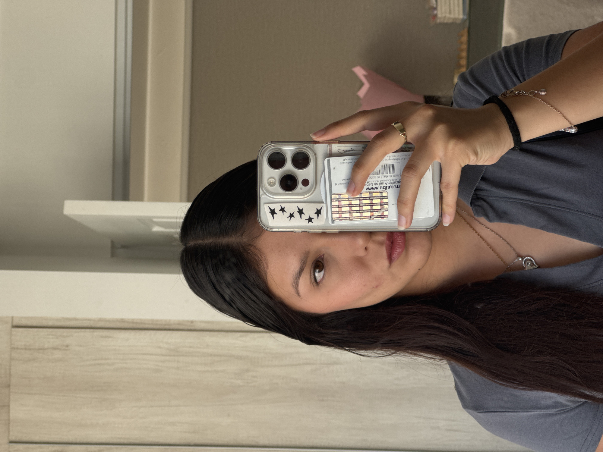 A woman is taking a selfie with her phone. The phone has a clear case with black star stickers on the bottom and a credit card or ID tucked behind it. She is wearing a gray t-shirt, a necklace with a heart-shaped pendant, a ring, and a bracelet. She has long dark hair. The background includes a wall, a white door, and a glimpse of something pink. The OCR text on the phone case indicates web addresses.