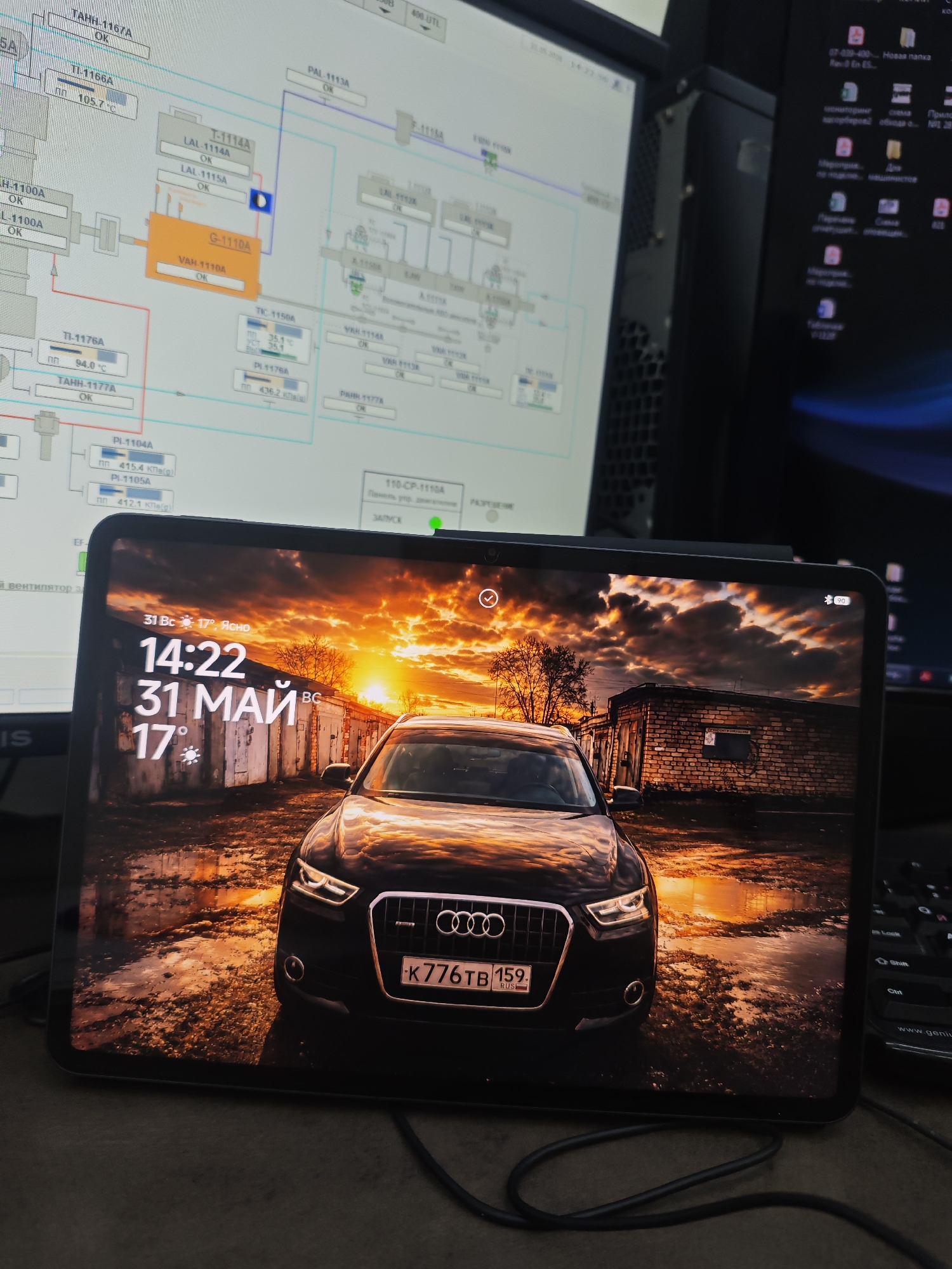 The image shows a tablet displaying a lock screen with a picture of a black Audi Q3 parked in front of a row of garages under a sunset or sunrise. The car's license plate reads "K776TB 159". The lock screen also displays the date and time as "31 MAY" and "14:22", with a temperature of "17°C". The tablet is placed on a desk near a computer monitor displaying diagrams and text, suggesting a technical or engineering environment. Another monitor to the right shows Windows desktop icons. A black cable connects to the tablet.