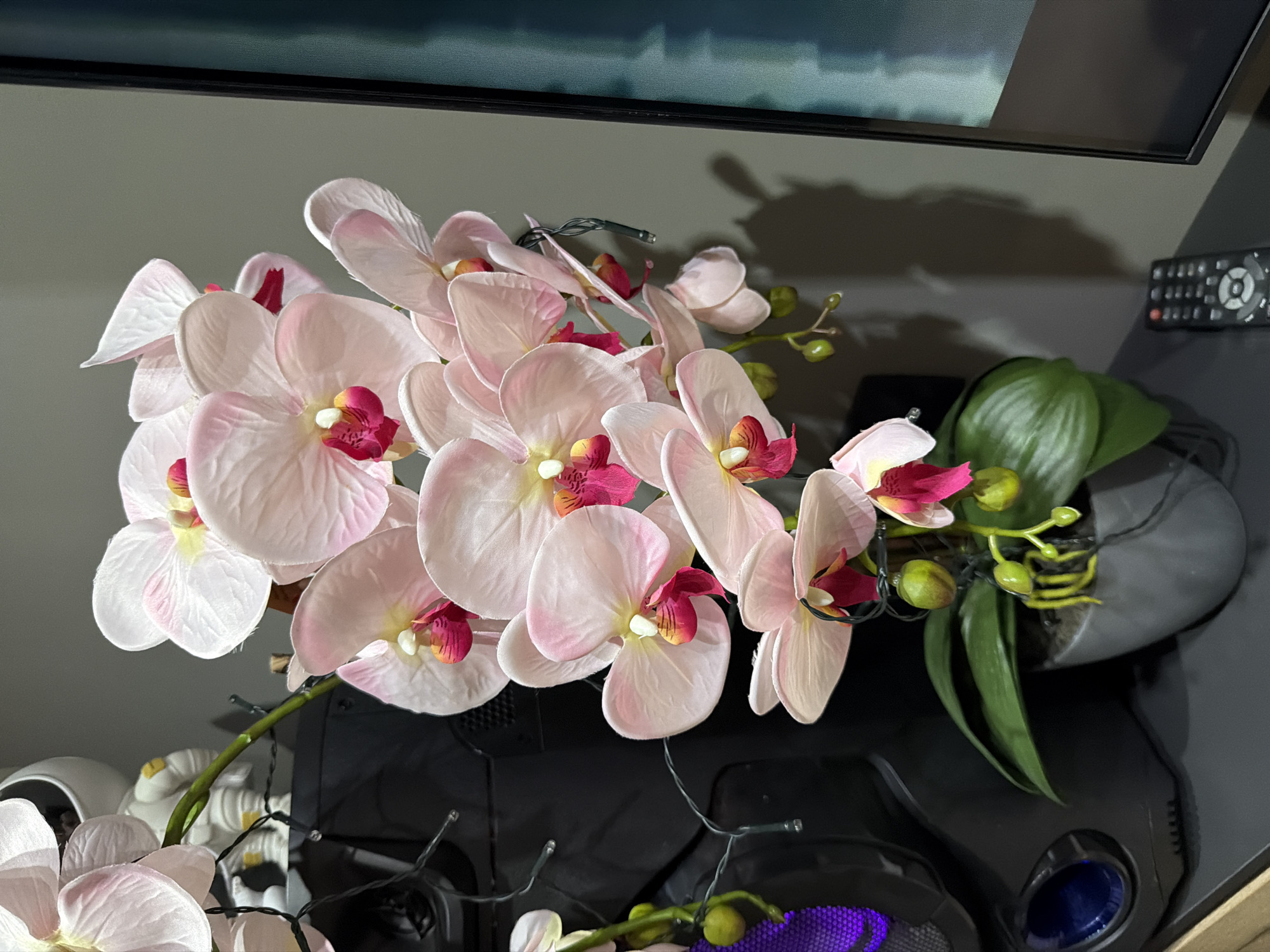 A close-up, eye-level shot captures a cluster of artificial light pink orchids with magenta centers, cascading from a pot. The orchids are adorned with a strand of small, clear fairy lights. To the left, a black computer tower with blue and purple lighting is partially visible. In the background, a white astronaut figurine stands on top of the computer tower. The orchids are placed on a dark, flat surface, likely a desk or shelf, with a gray wall behind them. A black remote control rests on the surface in the lower right corner. The overall lighting is dim, creating soft shadows. This scene is located in the city of Rafailovići, Montenegro.
