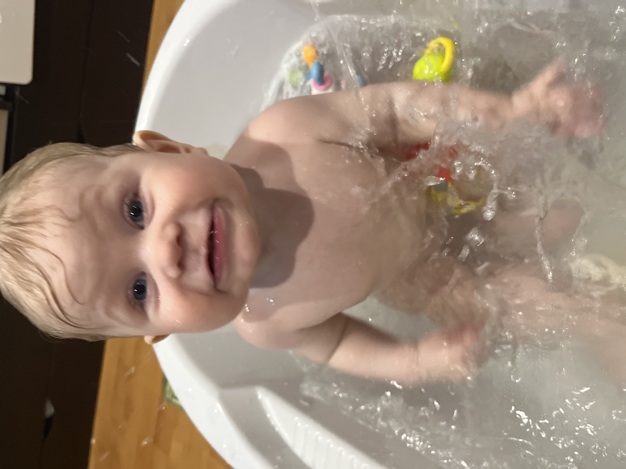 A baby is in a white bathtub filled with water. The baby has light blonde hair and blue eyes, and is looking up at the camera. There are several bath toys floating in the water. The background shows a wooden floor and a dark cabinet. There is no evidence to suggest that this image was taken in Vesyoloye, Russia.