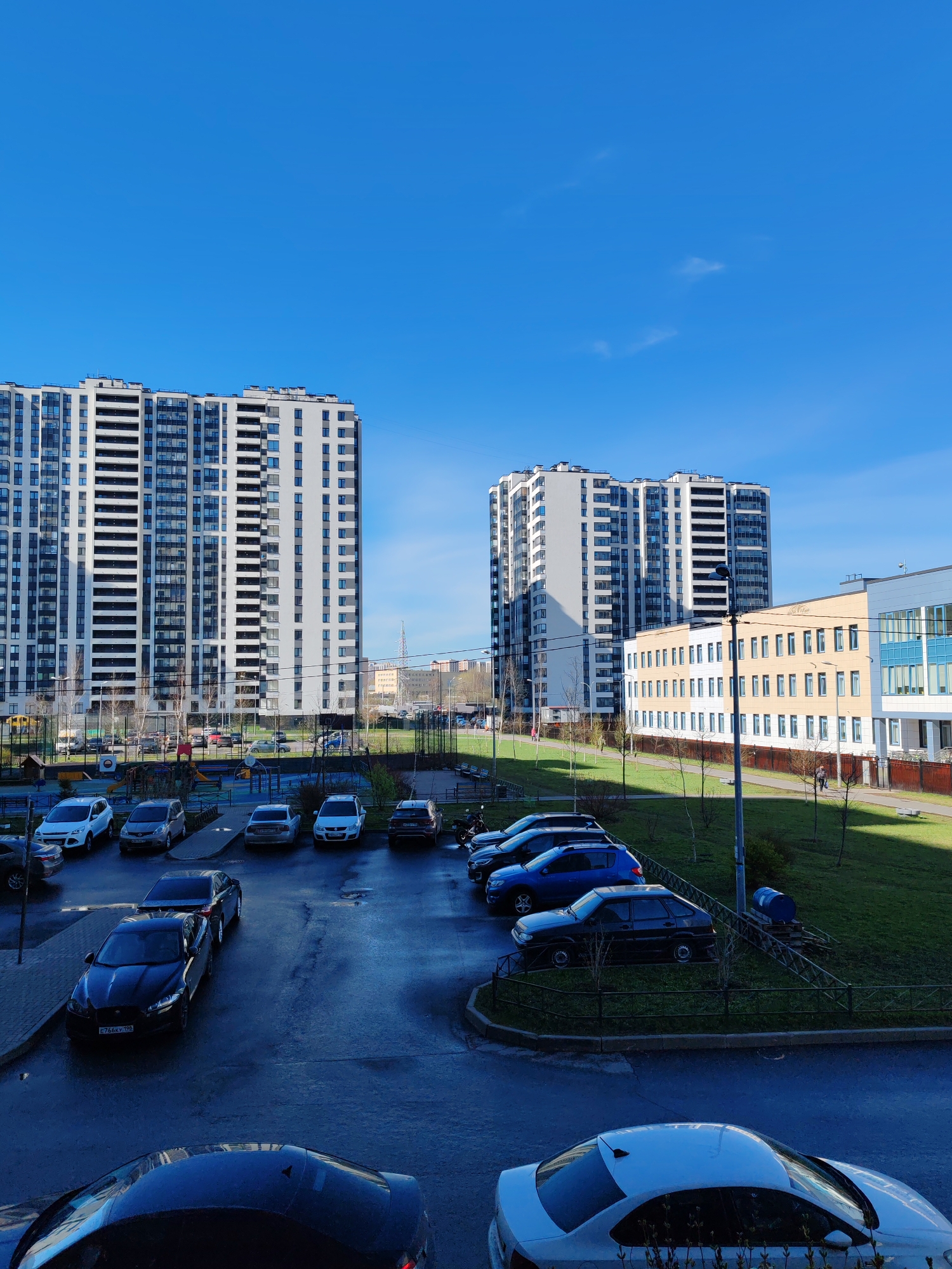 This media file captures an outdoor scene in a modern residential area of Saint Petersburg, Russia, under a clear, bright blue sky with a few wispy clouds. The time of day is sunny daylight, likely morning or early afternoon.
The primary subjects are two tall, contemporary apartment buildings with white and grey facades and numerous windows, standing prominently against the sky. To the right, a lower-rise building, possibly a school or educational institution, features sections of light yellow and light blue, with long rows of windows. Faint, indistinct text or a logo might be present above the windows of this building.
In the mid-ground and foreground, a paved parking lot is filled with various parked cars. Notable among them is a dark blue or black Jaguar sedan with the Russian license plate `E766KY 198` (indicating the Saint Petersburg region). Other sedans and SUVs are also visible. On a grassy bank to the right, an older, dark hatchback is parked unusually on an incline near a blue barrel, surrounded by a fence.
Green grassy areas with young, bare trees and a children's playground (featuring swings and slides) are scattered throughout the scene, indicating early spring or late autumn. A few small, indistinct human figures can be seen walking on paths near the buildings, suggesting typical daily urban activity. The overall impression is one of a calm, everyday urban environment.