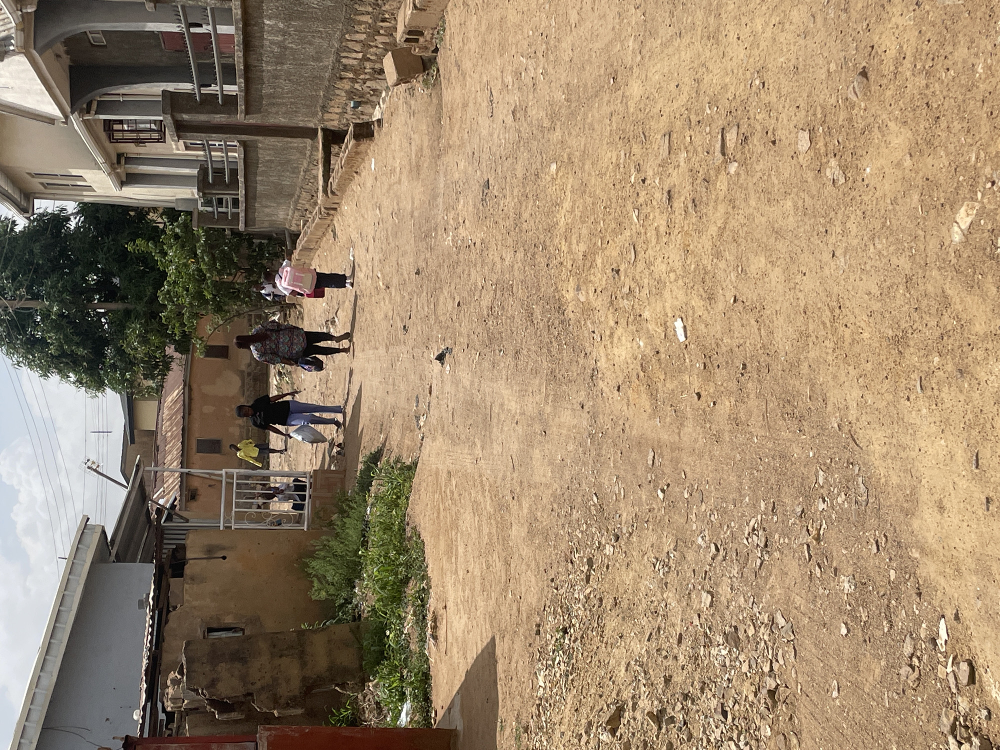 The image depicts an unpaved street in Jos, Nigeria. The ground is light brown dirt and gravel, with scattered debris. Several people are walking along the street. On the left side of the frame are various buildings, including some with corrugated metal roofing and others with concrete construction. Vegetation grows along the edges of the buildings. The sky is partially visible with light clouds.