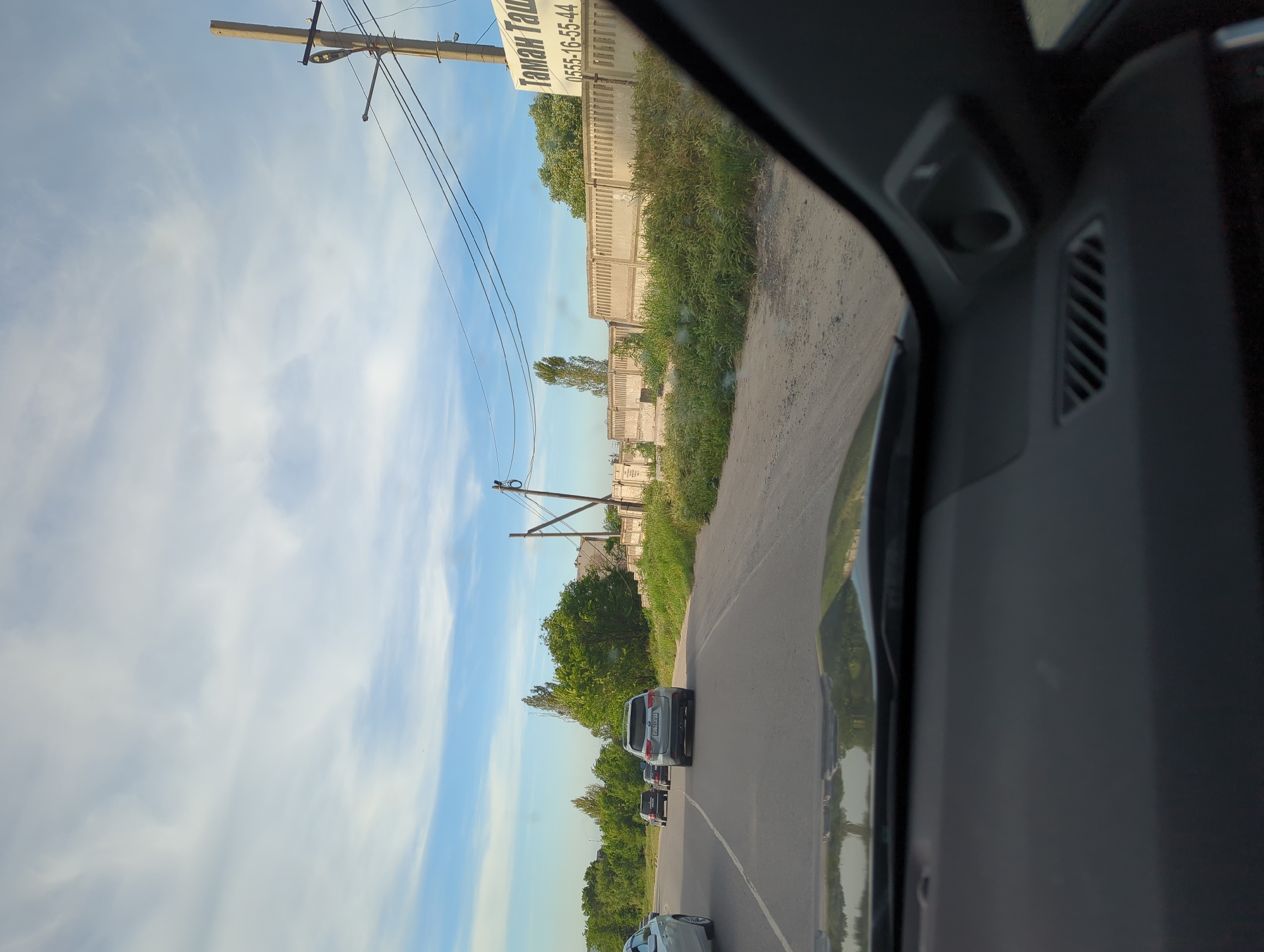 The image, apparently taken from inside a vehicle, depicts a road in Beş-Küŋgöy, Kyrgyzstan. The road is lined with vegetation on one side and a concrete wall with a sign in Russian on the other. Power lines run parallel to the road. Several vehicles are visible on the road, including at least one SUV. The sky is blue with scattered clouds. The interior of the vehicle from which the photo was taken is visible on the right side of the frame.