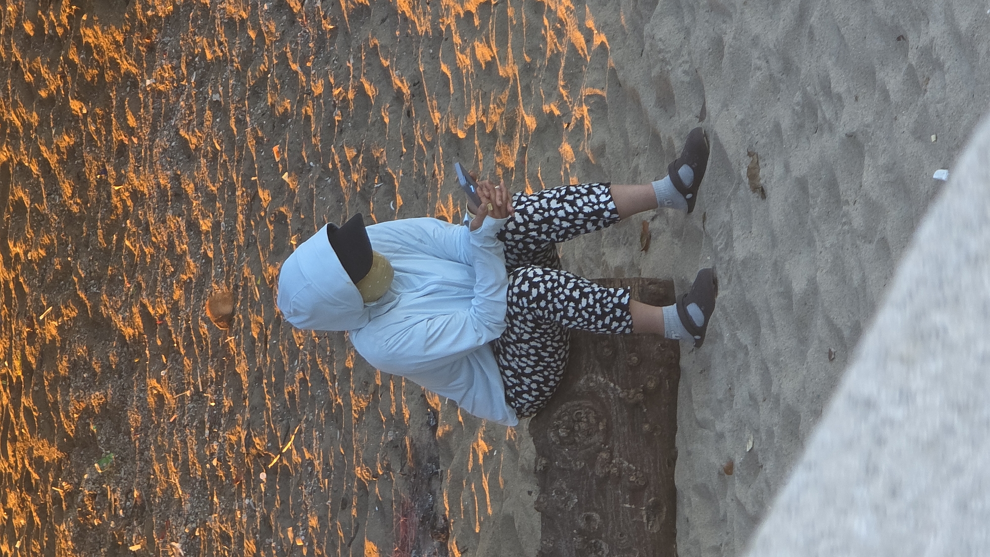 An individual is observed sitting on a large, dark brown log on a sandy beach in Nha Trang, Vietnam. The scene is illuminated by low-angle, warm, orange-toned light, suggesting either sunrise or sunset. The person is wearing a light blue hooded long-sleeve shirt, a dark baseball cap, and a yellowish fabric face mask that covers their lower face. They are dressed in black leggings with a white speckled or leopard-like pattern, grey ankle socks, and dark grey or black flat sandals. The individual's hands are clasped around a light blue smartphone, on which they appear to be focused. The immediate environment consists of sand textured by light and shadow, with scattered small debris including dried leaves and tiny pieces of what appears to be plastic or refuse. A light-colored concrete barrier or ledge is partially visible in the bottom left foreground.