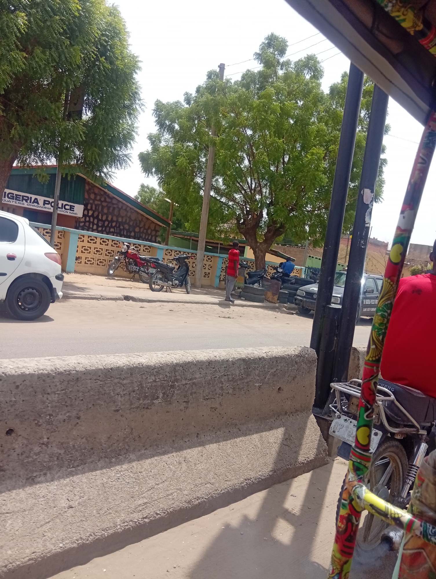 The image depicts a street scene in Gama, Nigeria. The view is from inside a vehicle, likely a motorized tricycle, indicated by the decorated frame on the right. In the background, a building identified as "Nigeria Police Headquarters Gwagwarwa Division Kano" is visible. Several motorcycles are parked near the building. A man in a red shirt is standing on the street. A dark-colored vehicle and stacked tires are seen further down the street. A white car is parked on the left side of the road. Trees line the street, and overhead power lines are present. A concrete barrier runs along the side of the road.