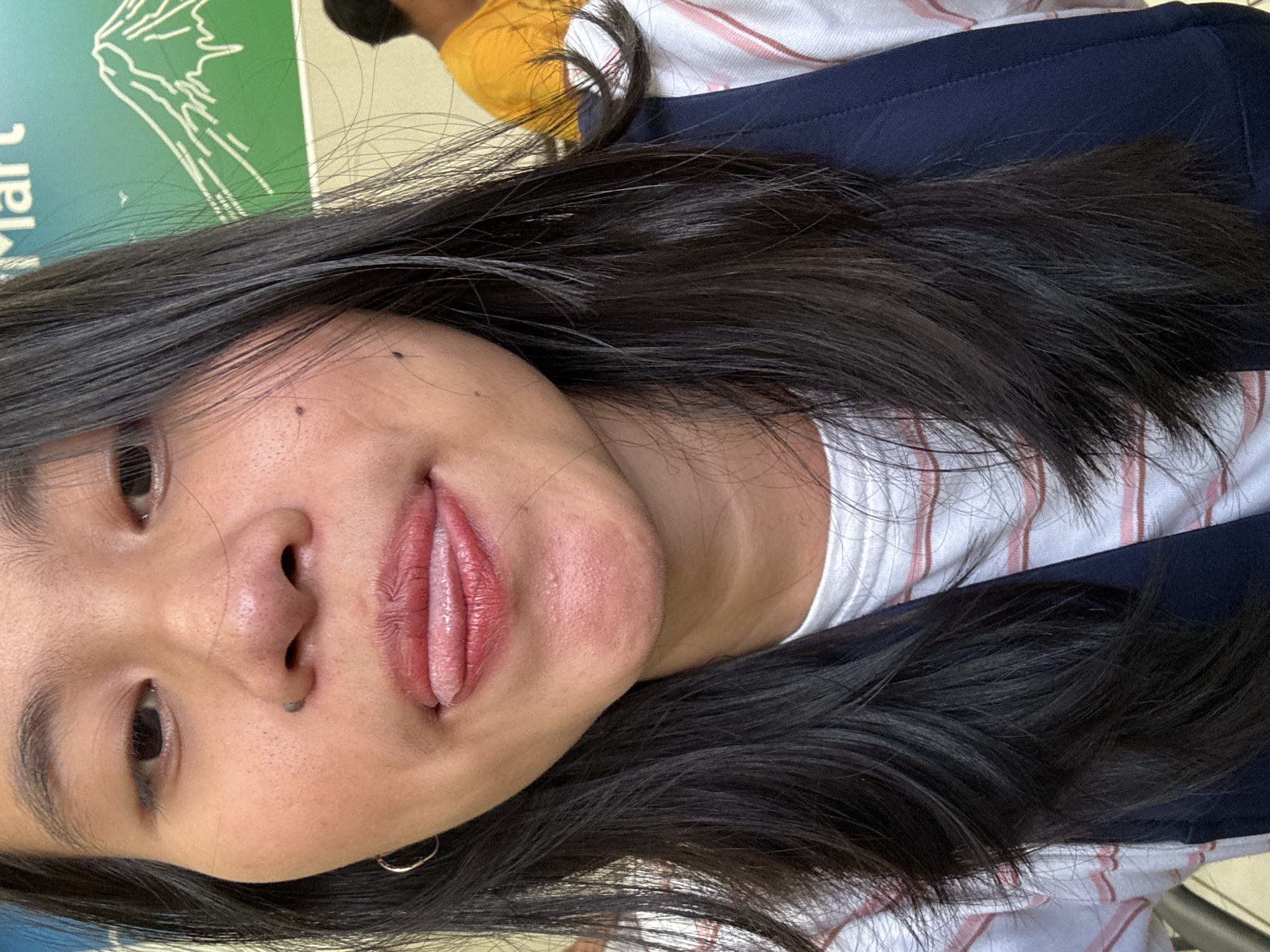 An extreme close-up selfie captures a young woman with long, dark hair in the foreground. She is sticking her tongue out, and her lips are adorned with reddish-pink lipstick. Her skin has visible pores and a small black piercing on her nose. A light hoop earring is visible on her left ear. She is wearing a white and pink striped t-shirt and a navy blue vest. In the background, a large green sign with a white outline drawing of a mountain and the text "Mart" is visible. A person wearing a yellow shirt is partially visible to the right of the frame. The image appears to be taken outdoors, possibly in a public space with some natural light.