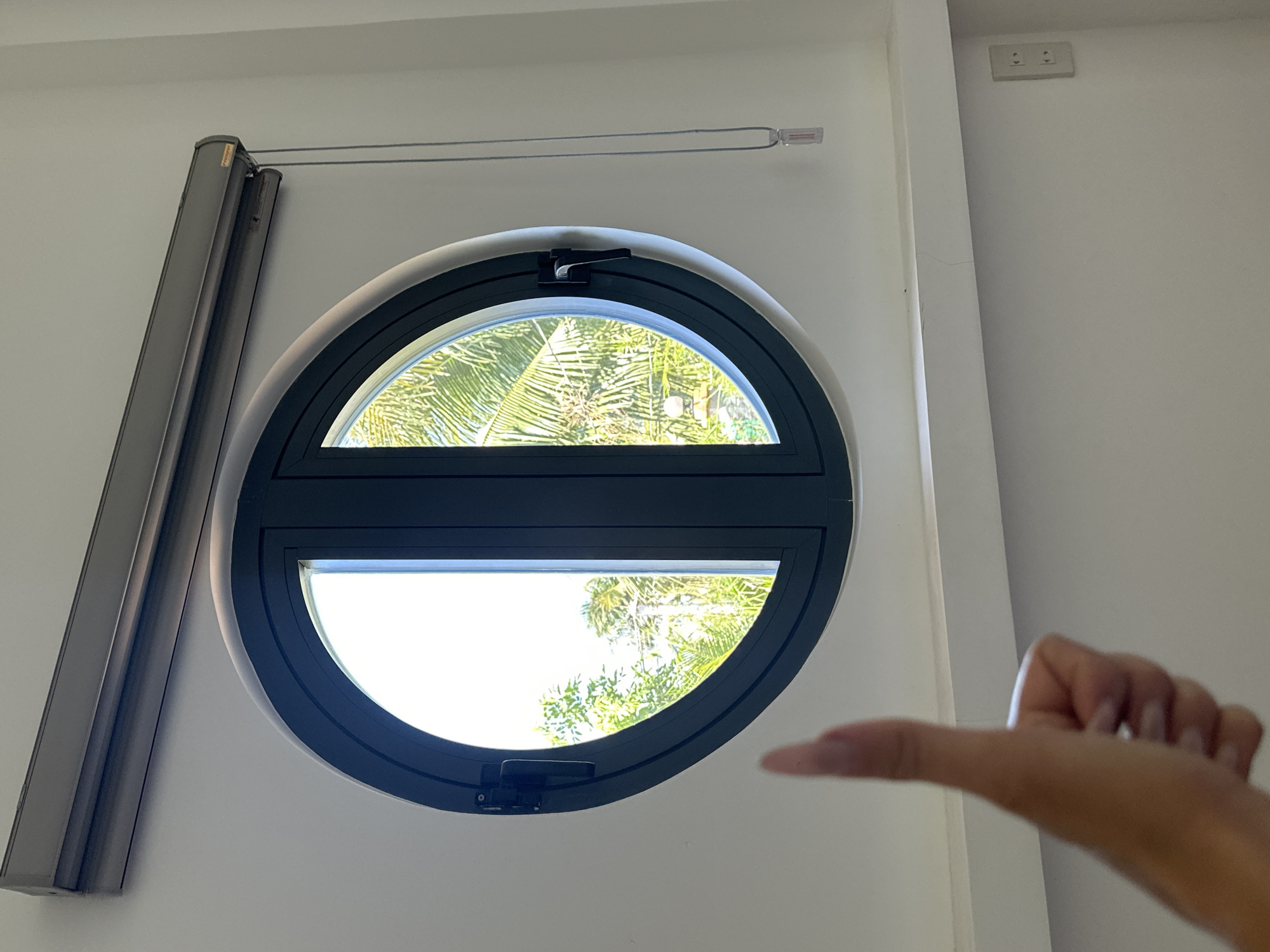 Here is a detailed description of the image:
The image is taken indoors, with a white wall as the backdrop. The focal point is a round window, bisected horizontally by a dark grey frame. The upper half of the window shows the green fronds of a palm tree against a bright sky. The lower half of the window also shows green vegetation, although the view is a bit brighter.
A gray shade system is fixed to the wall to the left of the window. To the right of the window is a partially visible hand, stretched out as if pointing.
In the top right corner of the image, there's a small white electrical outlet.
Based on the vegetation visible through the window, and the location name in the prompt, the location of this scene is in Mũi Né, Vietnam.