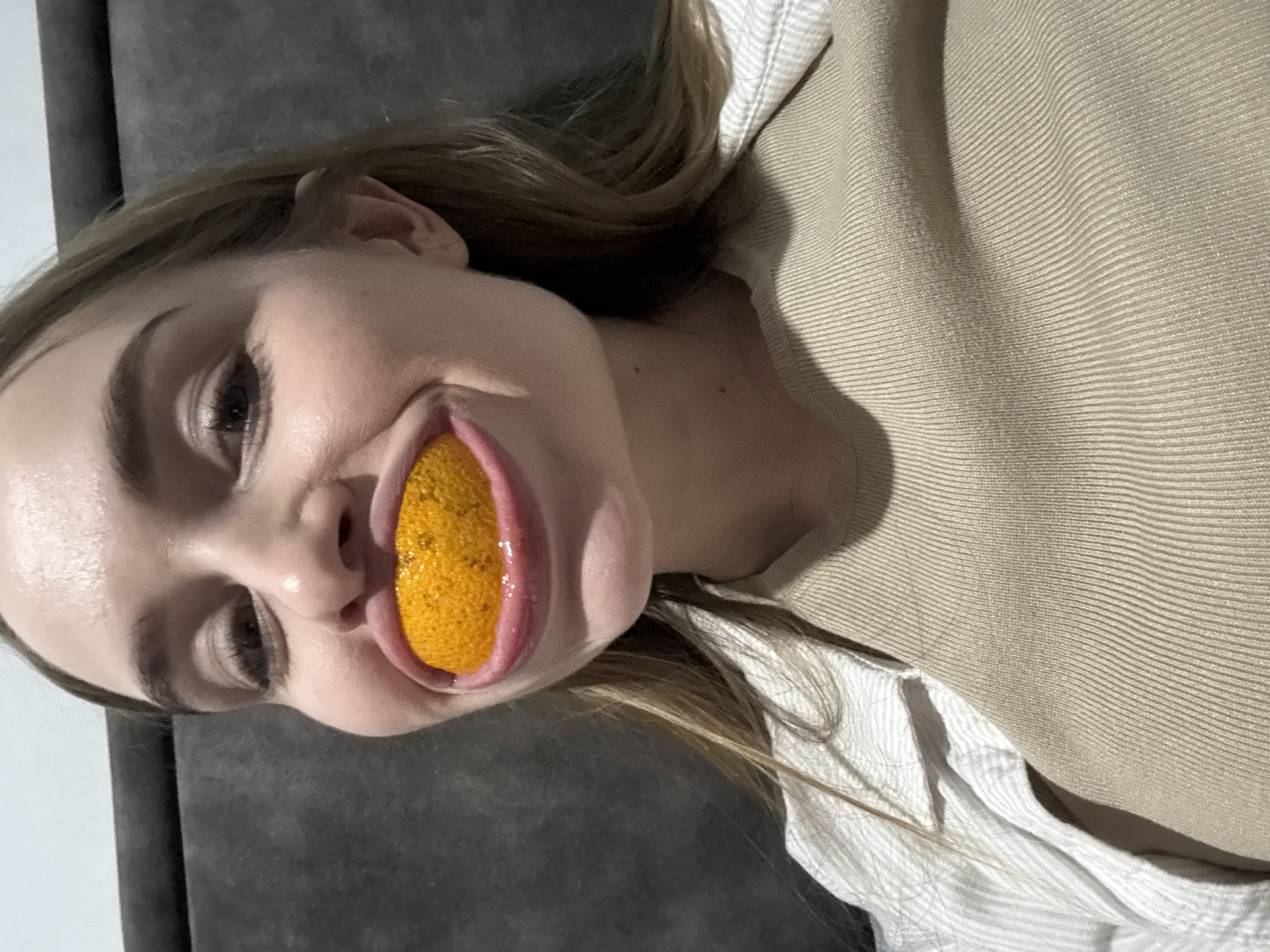 The image presents a close-up portrait of a female subject positioned indoors. Her face, from the forehead down to the upper chest, is visible, with eyes open and directed forward. A small, spherical, segmented orange object, consistent with a citrus fruit segment, is firmly held between her widely stretched lips, distorting their natural contour. The object appears moist with visible texture. She wears a beige, ribbed knit top layered under an open, light-colored, pinstriped outer garment. The background is an indistinct dark gray upholstered surface. The specific location within Budva, Montenegro, cannot be determined from the visual evidence in the image.