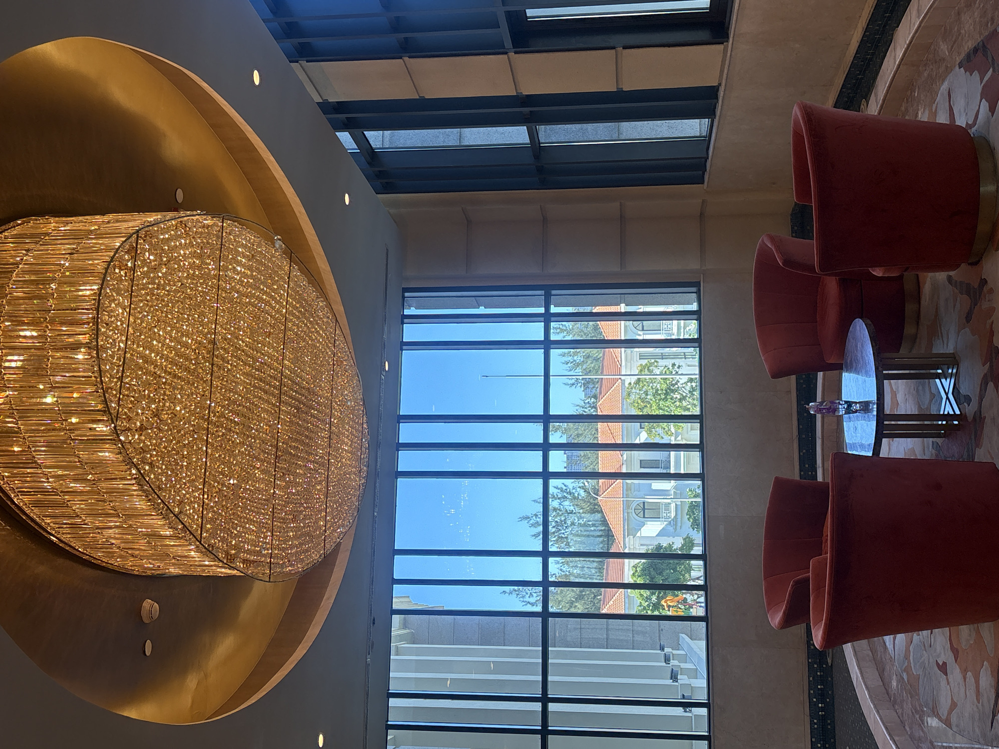 In a hotel lobby in Da Nang, Vietnam, a large, oval crystal chandelier hangs from a gold-accented ceiling. Below, a seating area is arranged around a small, dark-topped circular table. Four plush, red velvet armchairs are positioned facing inward, suggesting a conversation space. Through a large, grid-patterned window, a bright blue sky is visible, with white buildings and lush green trees in the distance. The architecture outside appears to be European-inspired, with red-tiled roofs. The floor features a patterned carpet in muted tones of beige and pink. The overall impression is one of elegant comfort and a welcoming atmosphere.