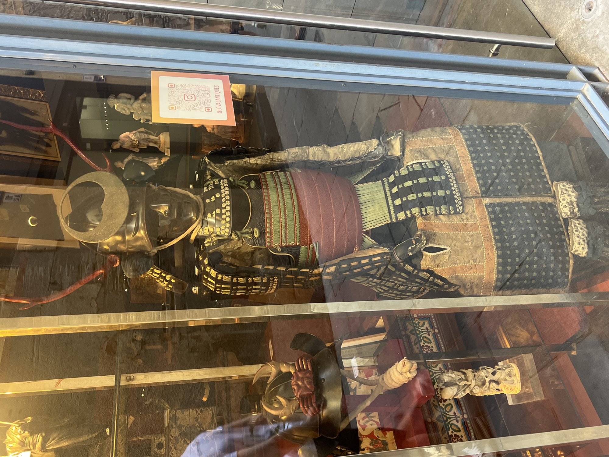 The image depicts a display window of an antique shop, likely located in Barcelona, Spain, given the architectural style visible through the glass. The main subject is a full suit of traditional Japanese samurai armor, displayed upright. The armor is detailed, featuring a metallic helmet with red accents and a facemask. The torso is protected by layered plating and a prominent reddish sash. The lower body is covered by armored skirts and leggings, elaborately decorated with patterns.
In the background, visible through the glass and reflections, are other antique items. These include figurines of people, decorative frames, and possibly paintings or textiles. A QR code sign with an Instagram handle "@LOVALANTIQUES" is affixed to the inside of the window. The lighting suggests the photo was taken during the day, with indoor lighting also contributing to the reflections on the glass. The floor outside the shop appears to be paved with cobblestones or similar material, consistent with older European city streets. There are no identifiable people in clear view within the shop, though a blurred figure might be seen through the reflections on the left side of the armor. The overall presentation suggests a retail environment specializing in historical artifacts and collectibles.