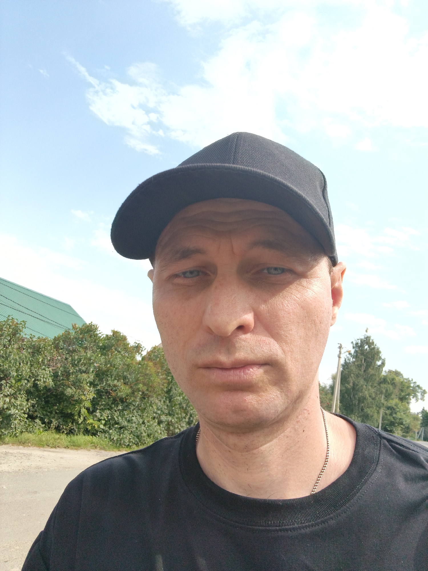 An outdoor, eye-level, selfie shows a man wearing a black baseball cap and a black t-shirt. He has light skin and blue eyes, and is looking directly at the camera. Behind him, a light green roof of a building is visible, as well as a line of trees and a blue sky with some clouds. The man is wearing a silver chain around his neck. He appears to be standing on a dirt road or path. This scene is located in Mokshan, Russia.