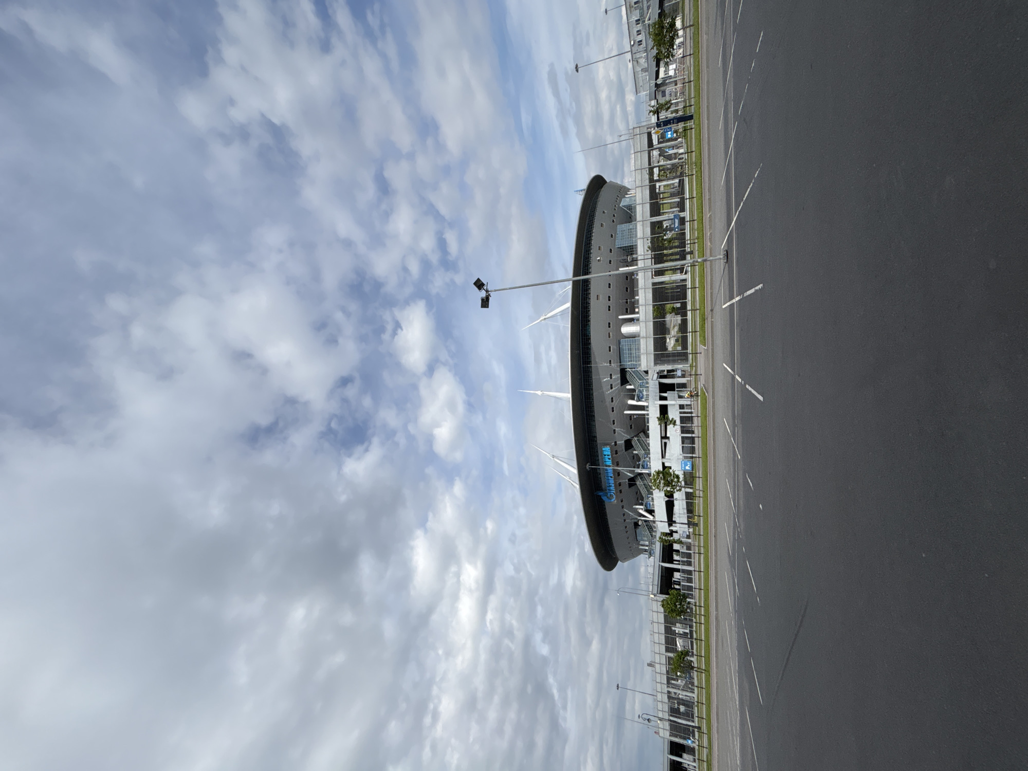 The image captures a large, modern stadium building under a partly cloudy sky, with an empty asphalt parking lot or access road in the foreground. The stadium, identified as Gazprom Arena through visible text, is an imposing structure with a curved, dark grey facade and a retractable roof design, featuring prominent white mast-like elements extending upwards. Large blue lettering on the stadium's side reads "GAZPROM ARENA" and possibly "ЗЕНИТ" (Zenit), indicating its association with the FC Zenit Saint Petersburg football club, which aligns with the Saint Petersburg, Russia context.
The foreground consists of a wide, dark asphalt surface, marked with white dashed lines for parking or lanes, suggesting a large capacity area usually bustling with activity, but currently devoid of vehicles or people. Light poles with multiple floodlights are sparsely distributed across this area and near the stadium. A metal fence runs along the edge of the asphalt, separating it from the stadium complex, which includes some sparse trees and shrubs.
The weather appears to be daytime, with a bright sky featuring a mix of grey and white clouds, suggesting an overcast or partly cloudy day. No specific events or activities are taking place, creating a calm and empty atmosphere around the massive stadium.