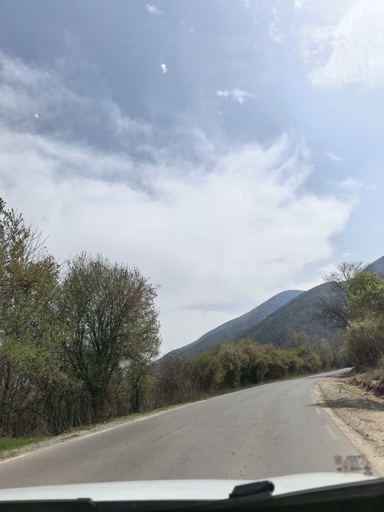 A daytime shot depicts a winding asphalt road flanked by trees and foliage, with a mountain range visible in the background under a partly cloudy sky. The photo appears to be taken from the perspective of someone in a vehicle, as part of the dashboard is visible at the bottom. The location is in the city of Unknown, Unknown.