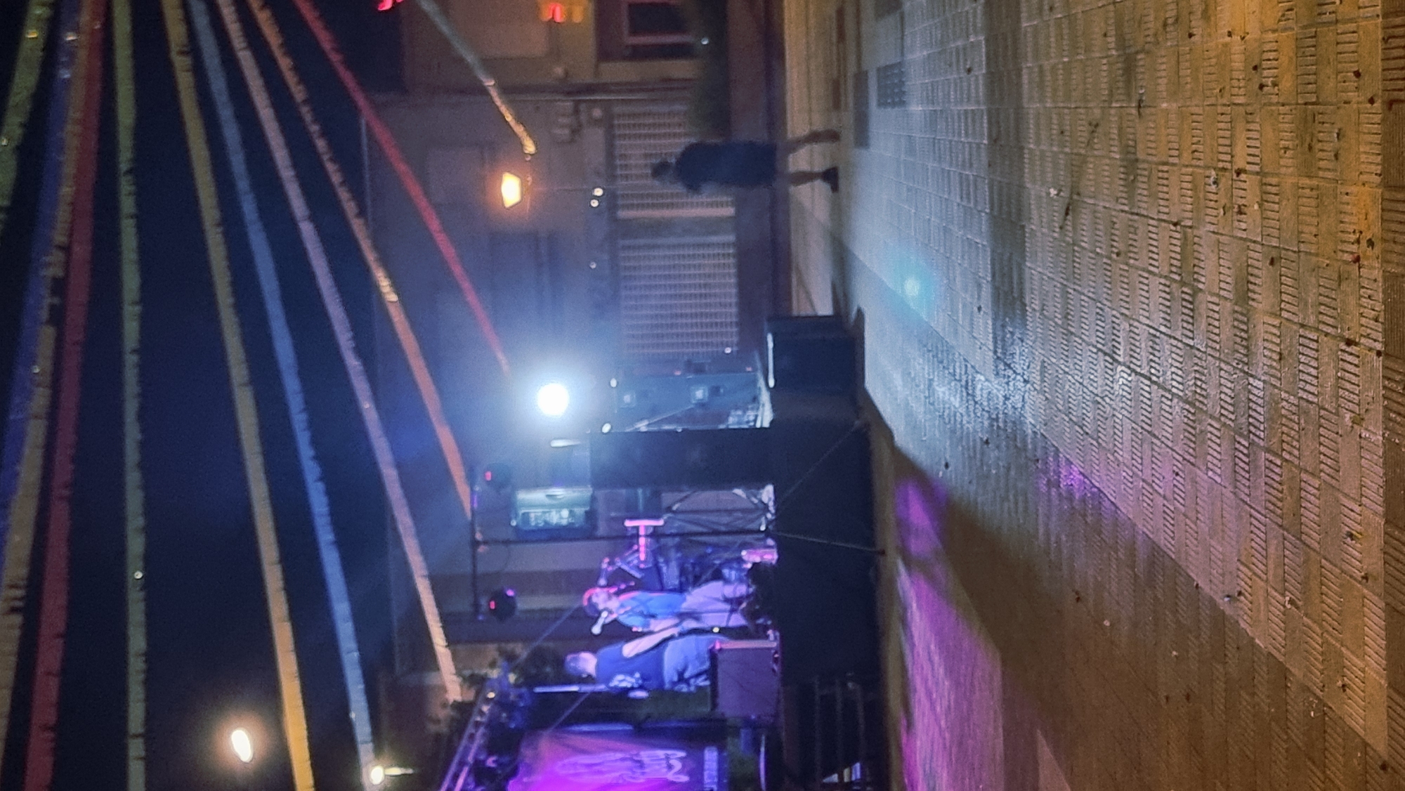 A nighttime outdoor scene in Sant Joan Les Fonts, Spain, features a temporary stage set up for a performance. Two individuals are visible on the stage: one in a blue shirt and light-colored pants, and another in a dark t-shirt and shorts. They appear to be interacting with musical equipment. Behind them, a banner with an illegible logo hangs. Stage lighting illuminates the performers and the immediate area. In the background, colorful string lights are draped across the sky. A single person, silhouetted and facing away from the camera, walks on the paved ground in front of the stage. The ground is tiled with a pattern of raised rectangular elements, and it appears to be damp, reflecting the lights. Buildings and streetlights are visible in the distance.
