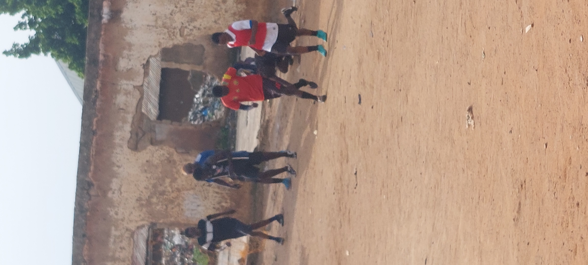 The image shows a group of people running in a dirt area. The scene is outdoors, with a dilapidated building in the background. The building appears to be made of mud or adobe, with a corrugated metal roof. There is also a pile of trash visible near the building.
There are at least five people in the image, all appearing to be young men, wearing athletic clothing. One person is wearing a red and white striped shirt, another a red shirt with yellow accents, and two are wearing black shirts with blue accents. The individuals are in motion, suggesting they are running, possibly as part of a race or training exercise. Their running posture and the dust kicked up by their feet reinforce this activity.
The lighting suggests it is daytime, likely with overcast or hazy conditions, as there are no strong shadows. The overall environment appears to be a rural or semi-urban area in Bukuru, Nigeria, given the building style and the unpaved ground. There is no visible text in the image. The atmosphere appears to be one of exertion and possibly friendly competition, as there are no overt signs of distress or aggression. The presence of the trash pile and the state of the building suggest an area that may lack robust waste management and upkeep.