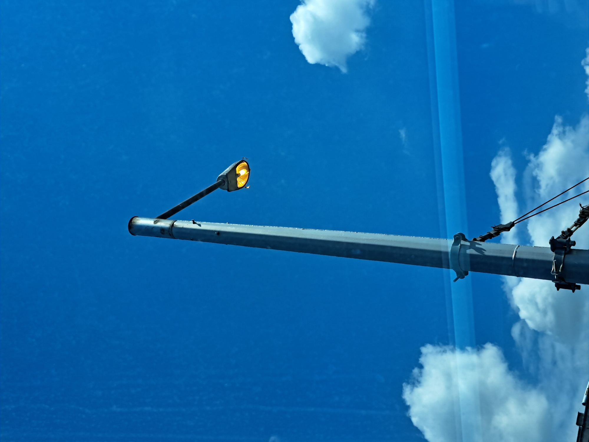 A low-angle shot captures a grey metal light pole extending from right to left across a vibrant blue sky, partially obscured by reflections of clouds and what appears to be a vertical structure on the right edge of the frame. The sky is a uniform blue on the left, transitioning to a sky filled with white clouds on the right, due to the reflection. The light fixture on the pole is illuminated with a yellow glow. The location cannot be determined with the information provided.