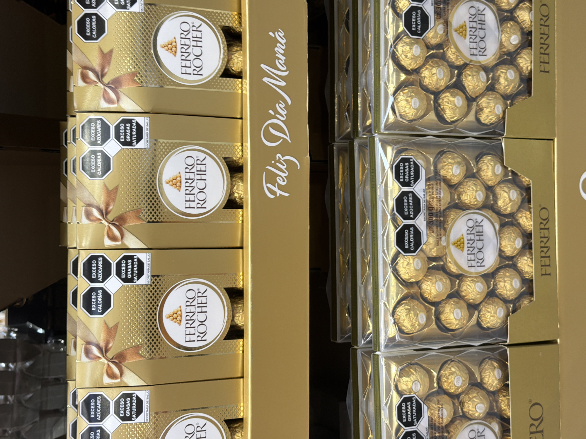 The image shows stacked boxes of Ferrero Rocher chocolates. Some boxes are gold with a brown bow and several warning labels. The text "Feliz Dia Mamá" is written in gold font on the right side of the boxes. The right side shows boxes of the same chocolate with the individual chocolate pieces visible through a clear window. The products are stacked and located in Santa Clara Ocoyucan, Mexico.