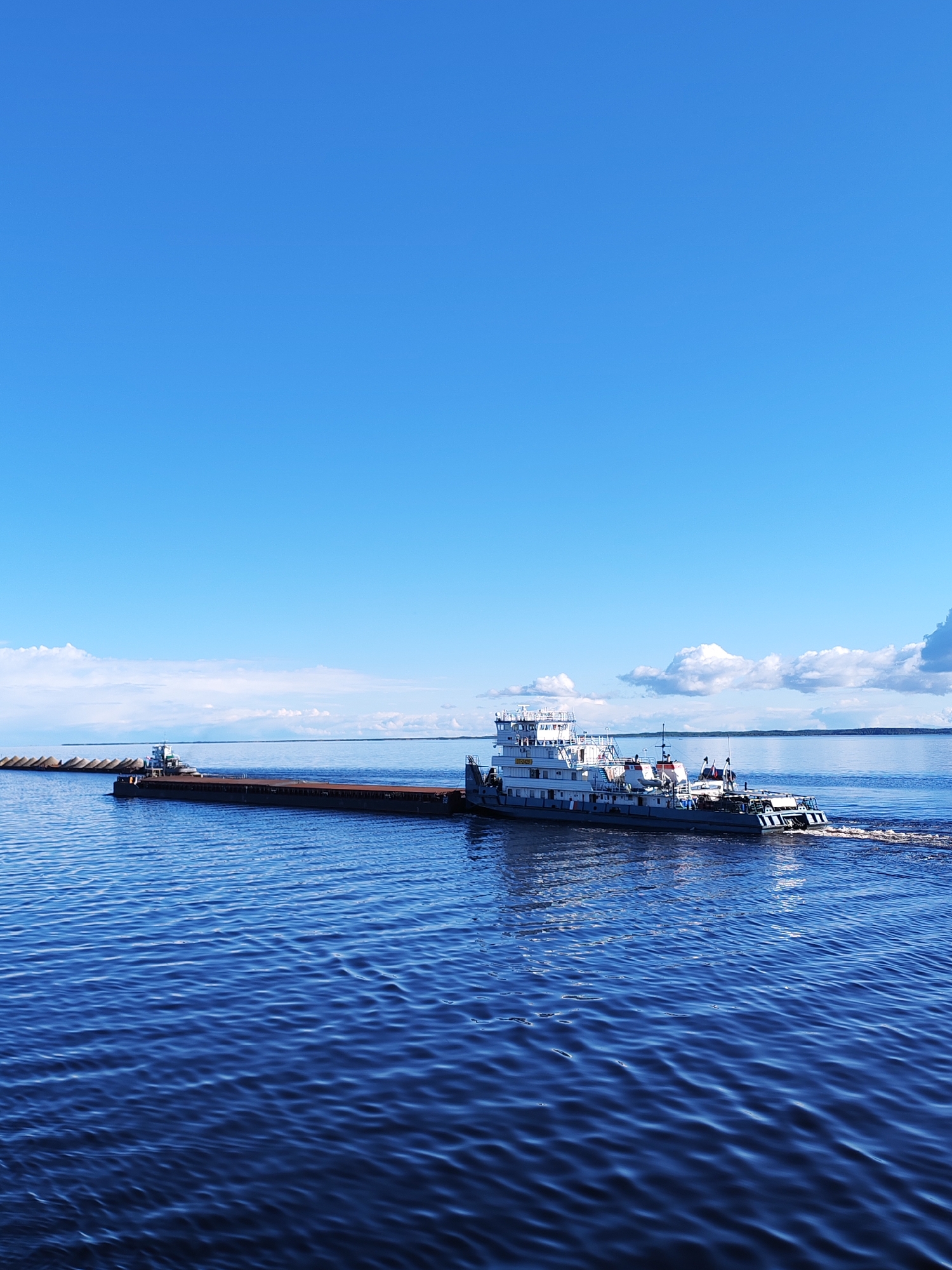 A wide-angle, outdoor shot depicts a tugboat pulling a long barge across a body of water, likely the Volga River near Mikhalevo, Russia, given the visual characteristics.
The tugboat is painted white with visible rust streaks. It is towing a long, dark-colored barge loaded with a cylindrical material. The barge also contains a crane near the far end of the shot. The tugboat is moving, as evidenced by the wake it creates in the water. The water is deep blue and rippled.
The sky is bright blue with a few scattered white clouds on the horizon. There is a faint treeline visible in the far distance.